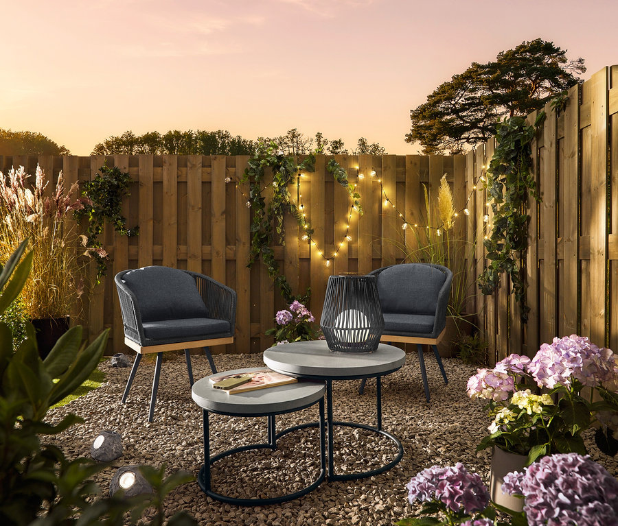 Zwei drehbare Gartenstühle mit Textil-Kreuzgeflecht stehen neben zwei Garten-Couchtischen mit Polyzement-Tischplatte. Eine kleine Solar-Laterne mit Textilband steht auf einem Tisch. 3 Solar-LED-Steine liegen im Kies.