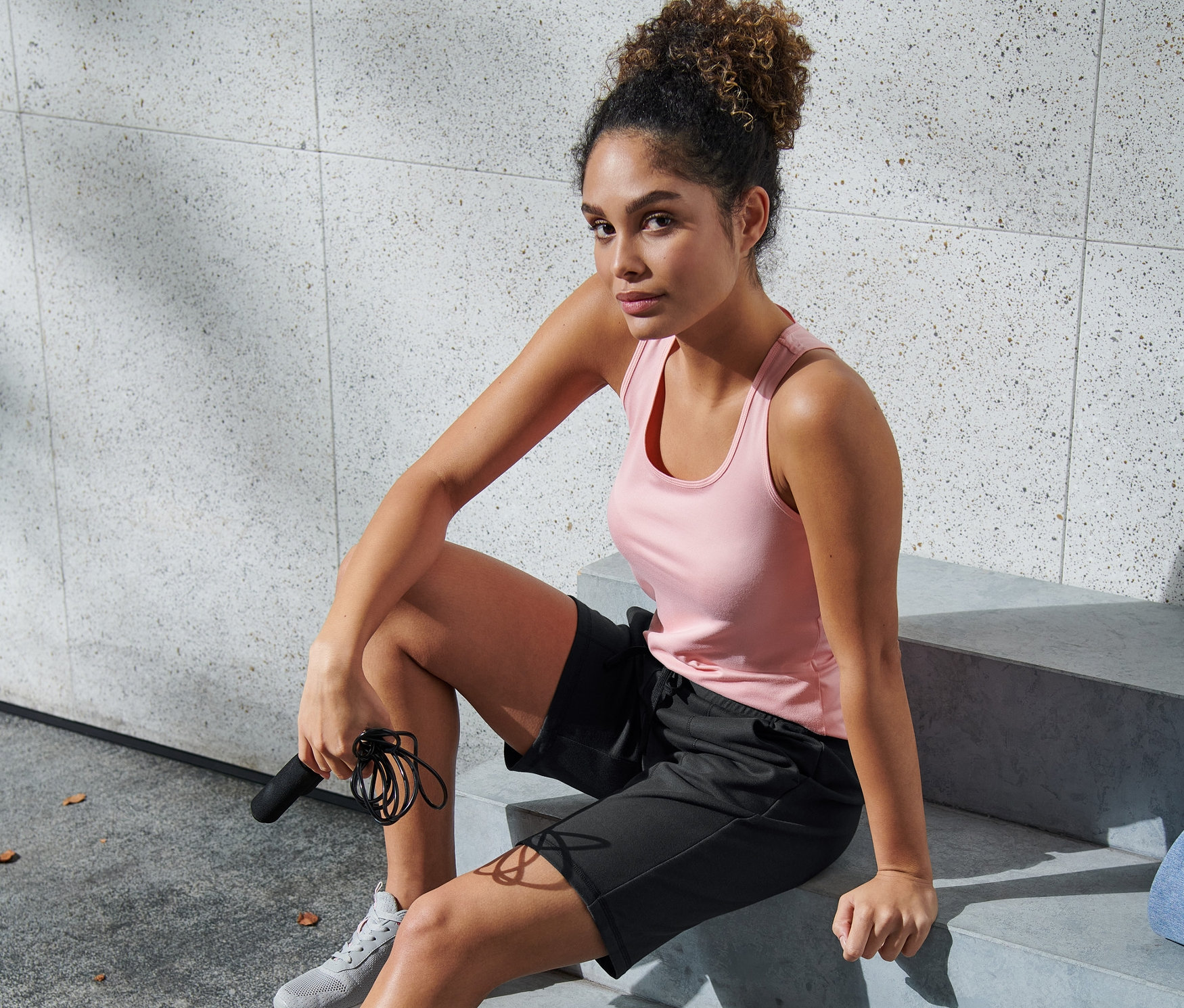 Frau sitzt in braun-schwarzen Funktionsshorts auf Treppe und hält Springseil.