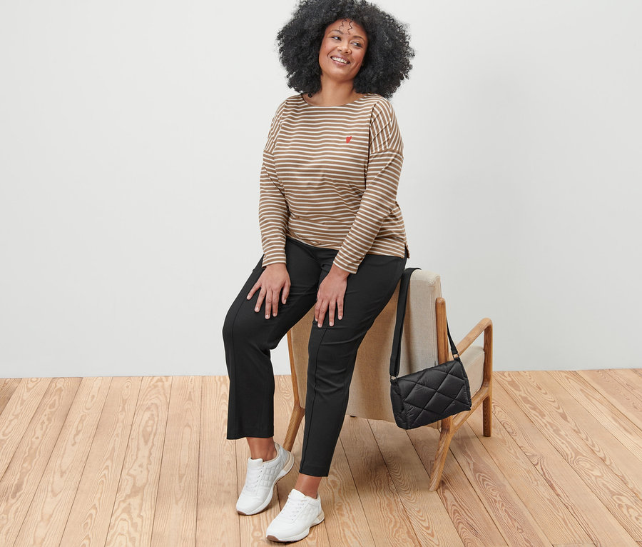 Frau sitzt auf einem Stuhl, trägt ein gestreiftes Langarmshirt, eine schwarze Sweathose und weiße Turnschuhe. Neben ihr befindet sich eine gesteppte Umhängetasche.