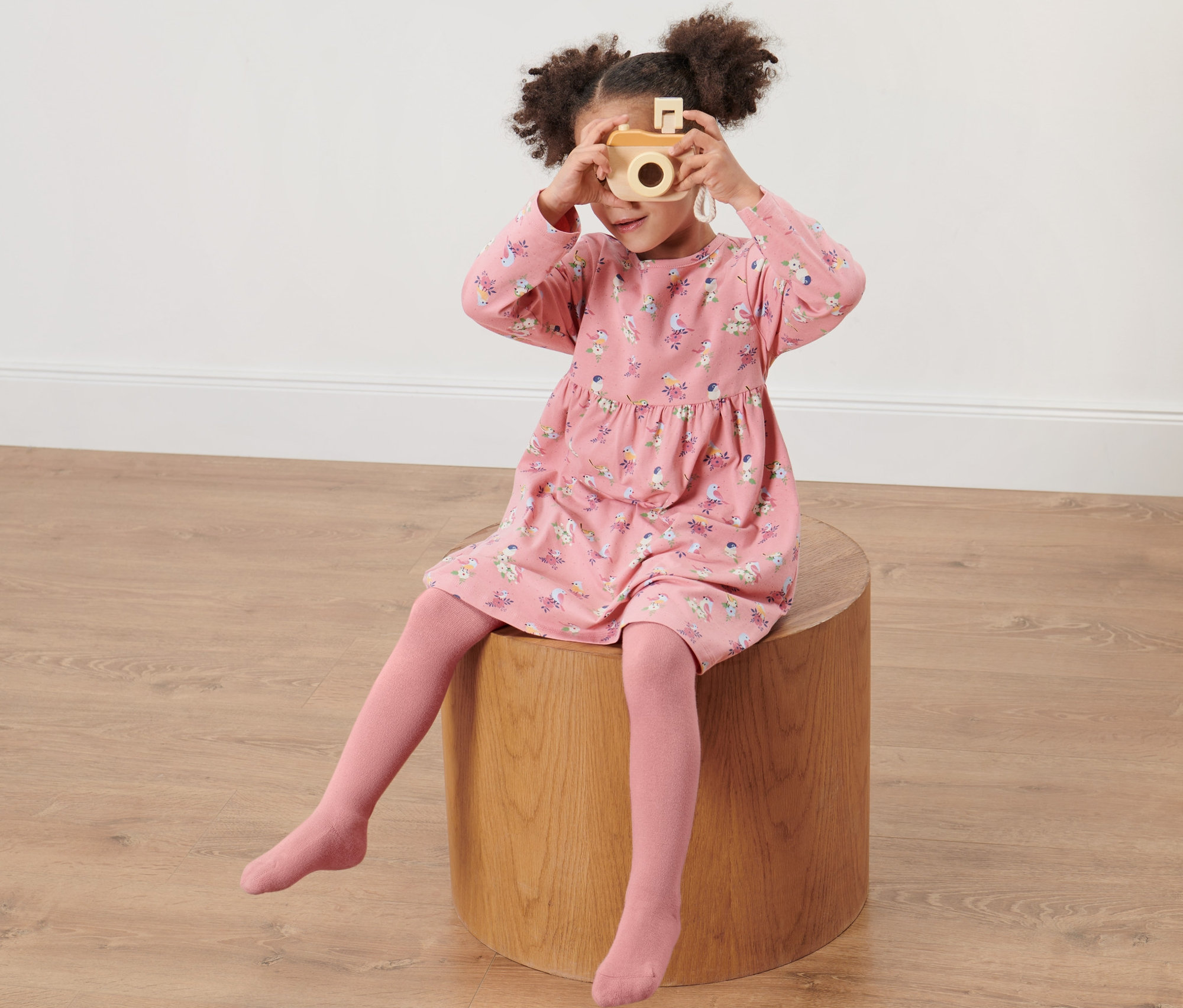 Mädchen sitzt auf einem Holzhocker und fotografiert mit einer Holzkamera. Sie trägt ein rosa Kleid mit Aufdruck und 2 Kinder-Strumpfhosen, rosa/hellblau.