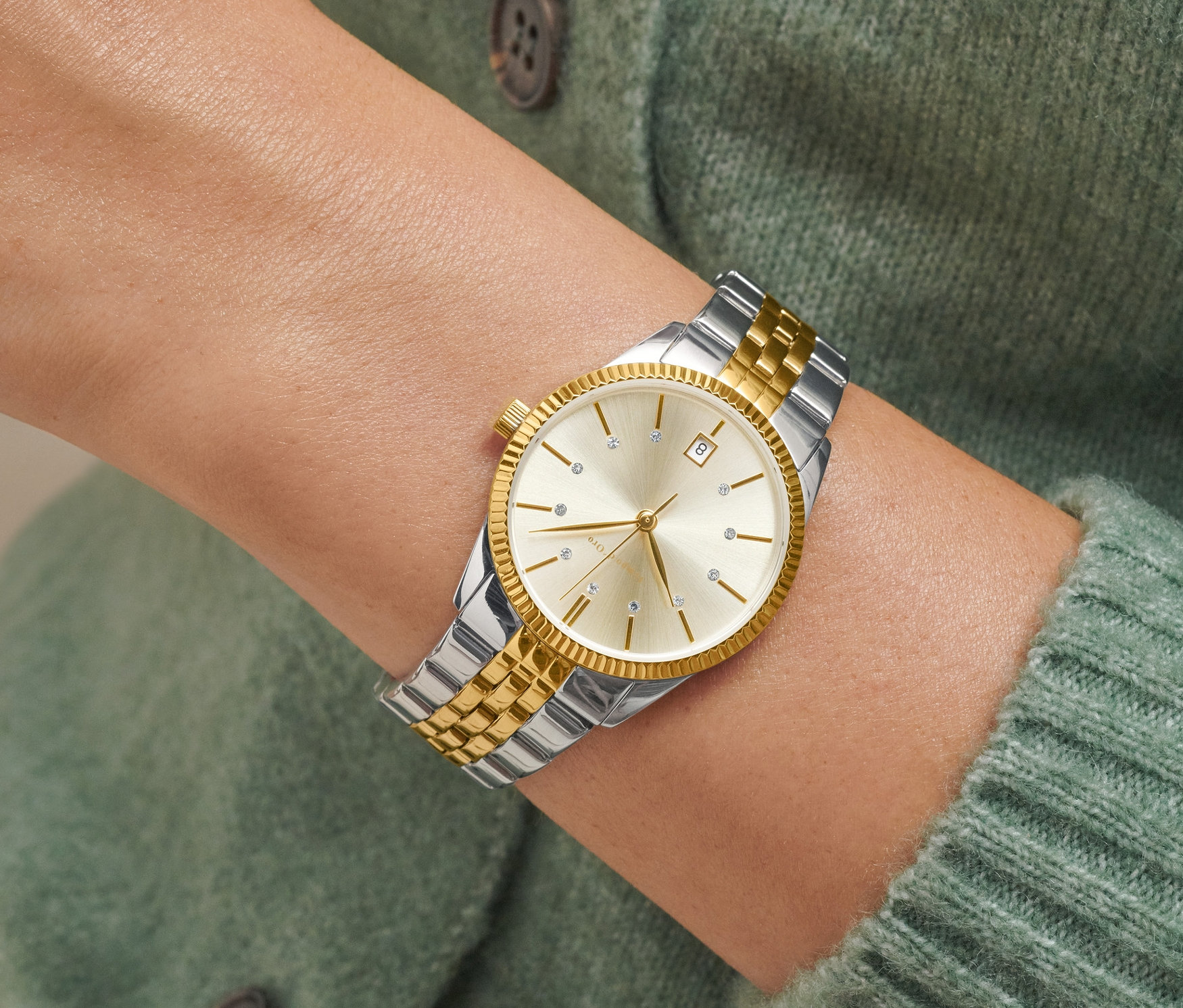 Detail einer Bicolor-Damenuhr mit Datum am Handgelenk einer Frau in einem grünen Pullover.