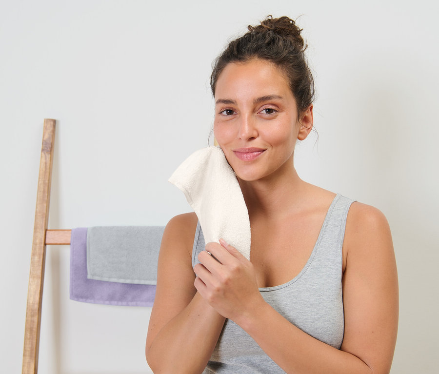 Frau trocknet ihr Gesicht mit einem weißen Handtuch ab. Dahinter befindet sich ein hölzerner Handtuchhalter mit einem grauen und einem violetten Handtuch.
