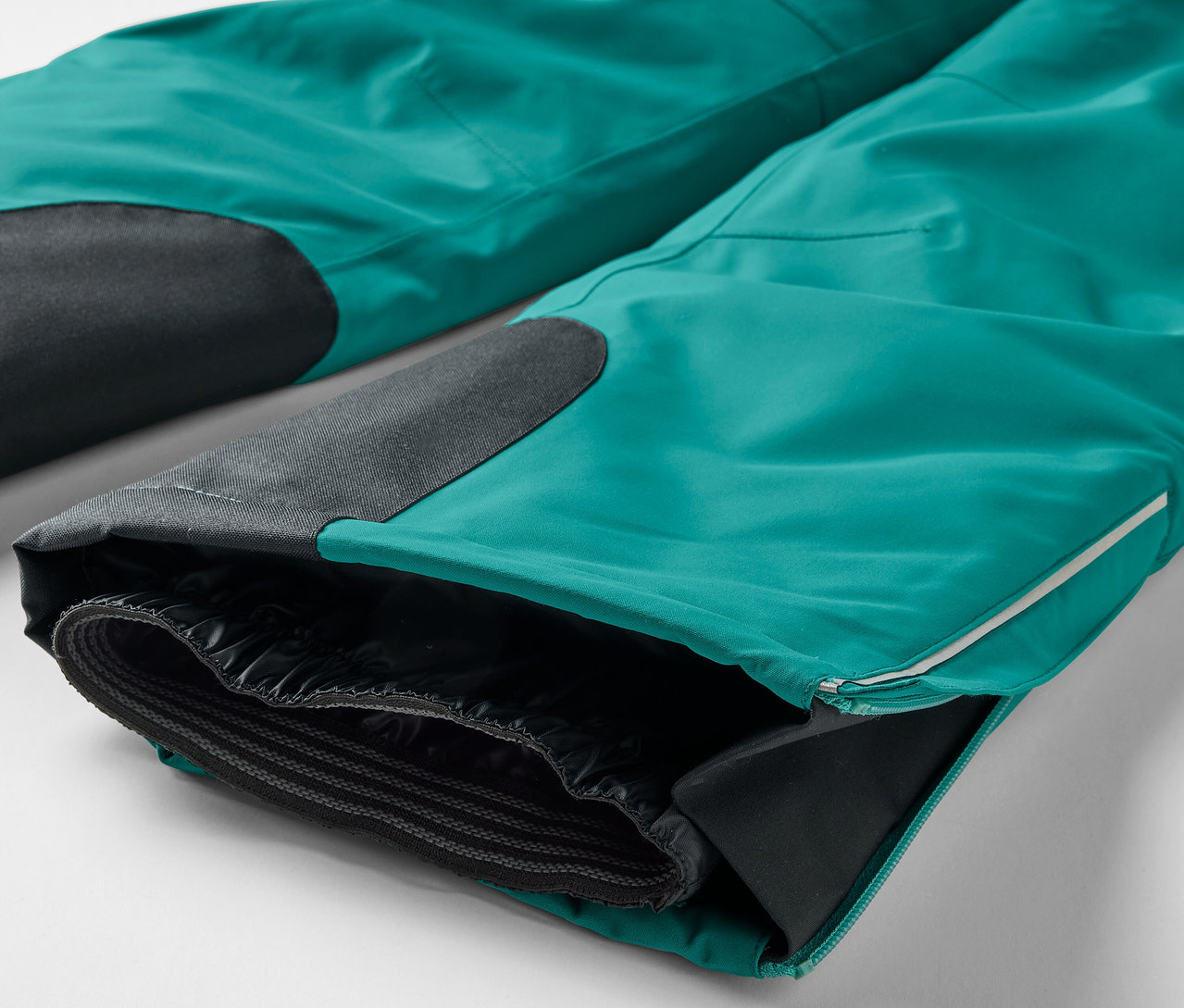 Detailaufnahme einer türkisfarbenen Hose mit schwarzen Kniepatches und einem geöffneten Hosenbeinsaum, der ein schwarzes Futter freilegt.