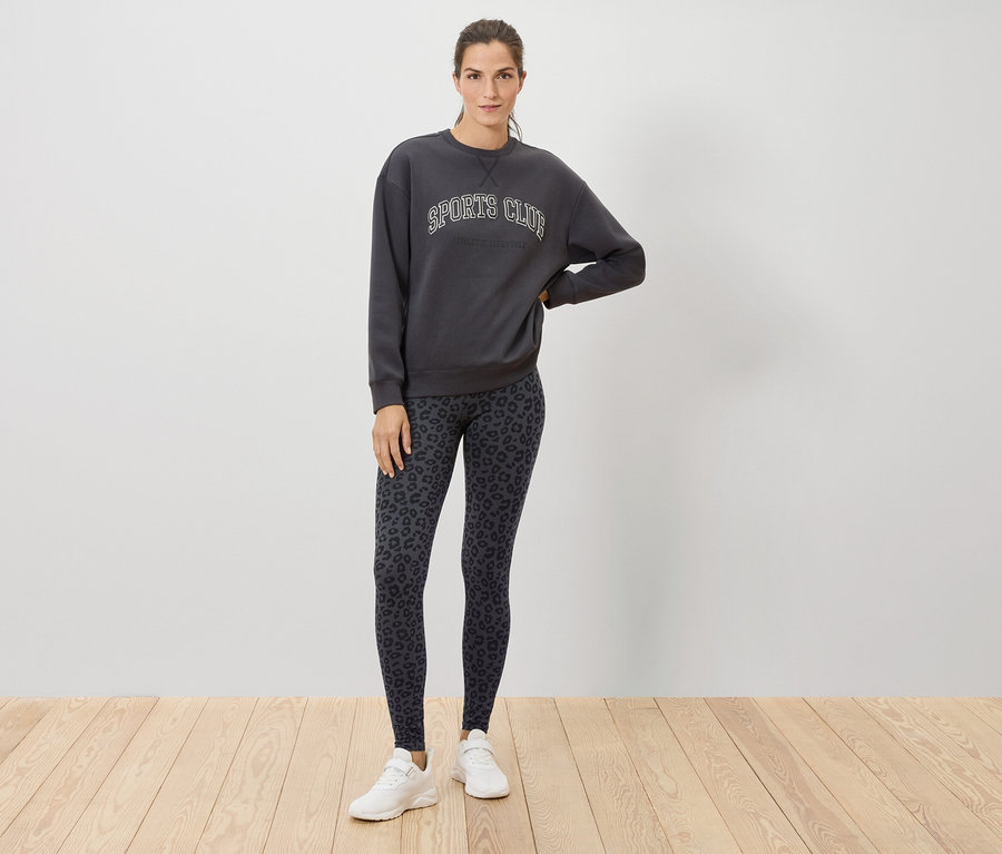 Eine Frau steht auf einem Holzboden und trägt ein graues Sweatshirt mit der Aufschrift SPORTS CLUB, Leggings mit Tiermuster und weiße Turnschuhe.