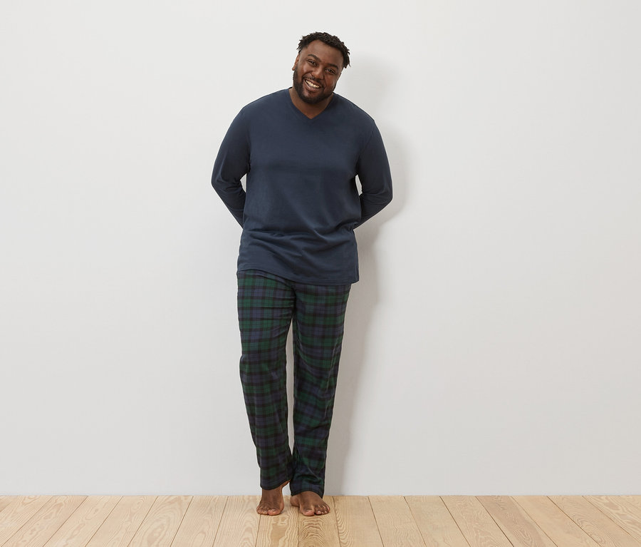 Ein dunkelhäutiger Mann steht da und lächelt in die Kamera. Er trägt ein dunkelblaues langärmeliges Schlafanzugoberteil und eine karierte Schlafanzughose.