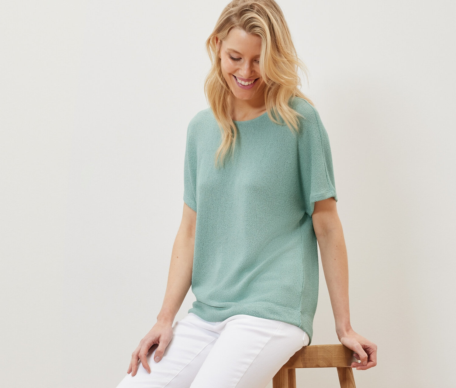 Blonde Frau sitzt auf einem Holzhocker und trägt ein strukturiertes Shirt in Salbei und weiße Hosen.