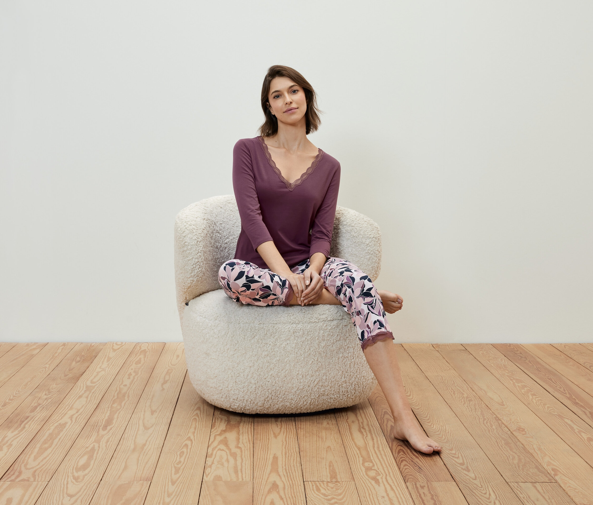 Eine Frau sitzt in einem Sessel und trägt ein violettes Shirt und eine geblümte Hose.