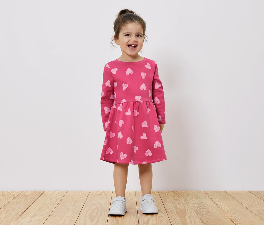 Ein lächelndes kleines Mädchen steht auf einem Holzboden und trägt ein rosa Kleid mit langen Ärmeln und Herzchenmuster.