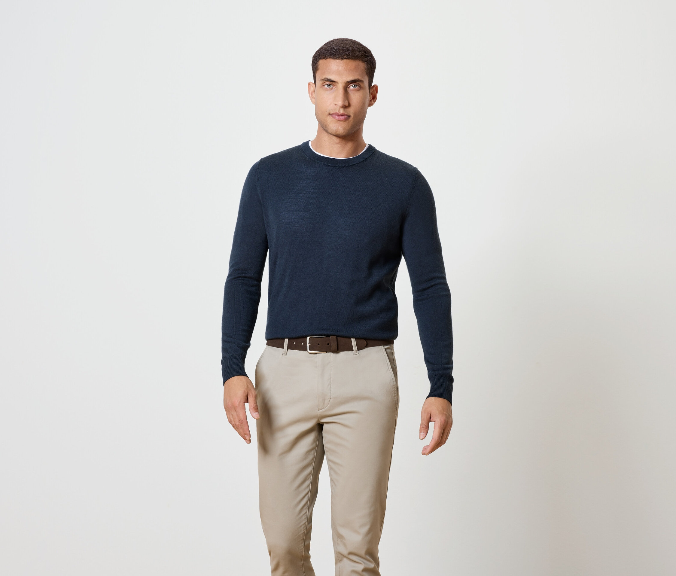 Mann steht und trägt einen dunkelblauen Pullover, einen braunen Gürtel und Khakihosen.