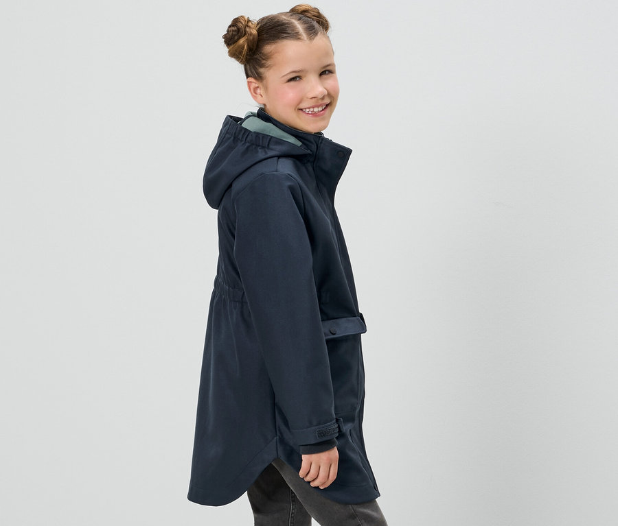 Lächelndes Mädchen posiert in marineblauem Kinder-Softshellmantel und grauen Jeans.
