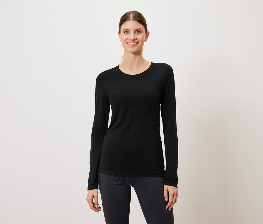 Frau trägt ein schwarzes Langarmshirt und dunkle Leggings vor einem weißen Hintergrund.