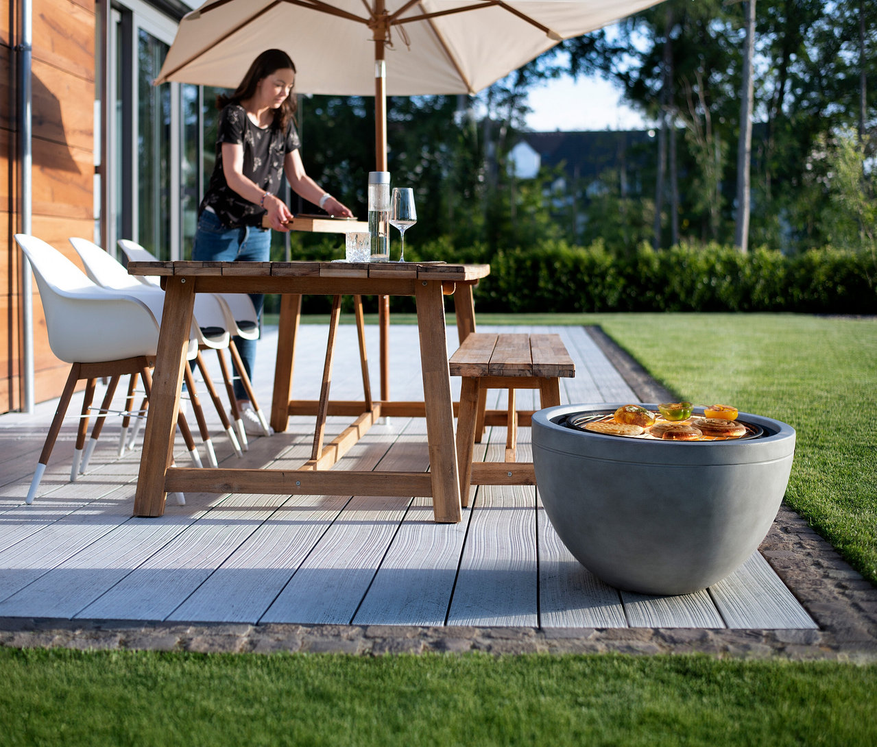 Eine Frau bereitet Essen auf einem Holztisch im Freien zu, mit einem grauen Grill im Vordergrund.