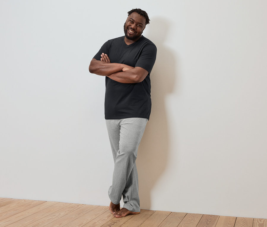 Ein dunkelhäutiger Mann mit verschränkten Armen lehnt an einer Wand, trägt ein schwarzes T-Shirt und graue Hosen.