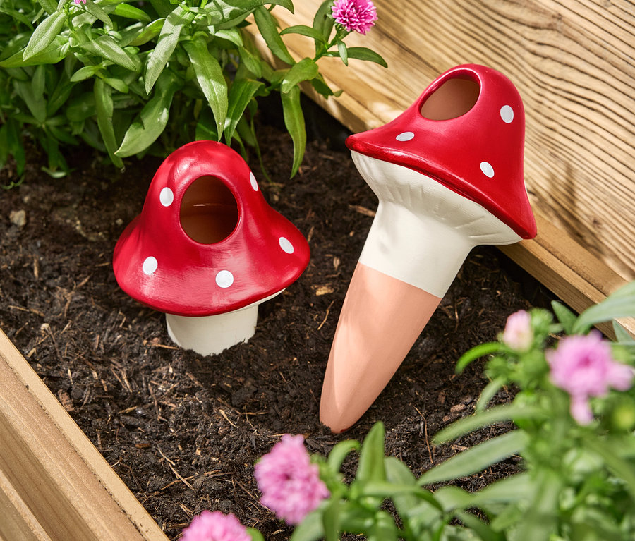 Zwei rote Pilz-förmige Bewässerungskugeln mit weißen Punkten stehen in einem Blumenkasten zwischen Blumen und Pflanzen.