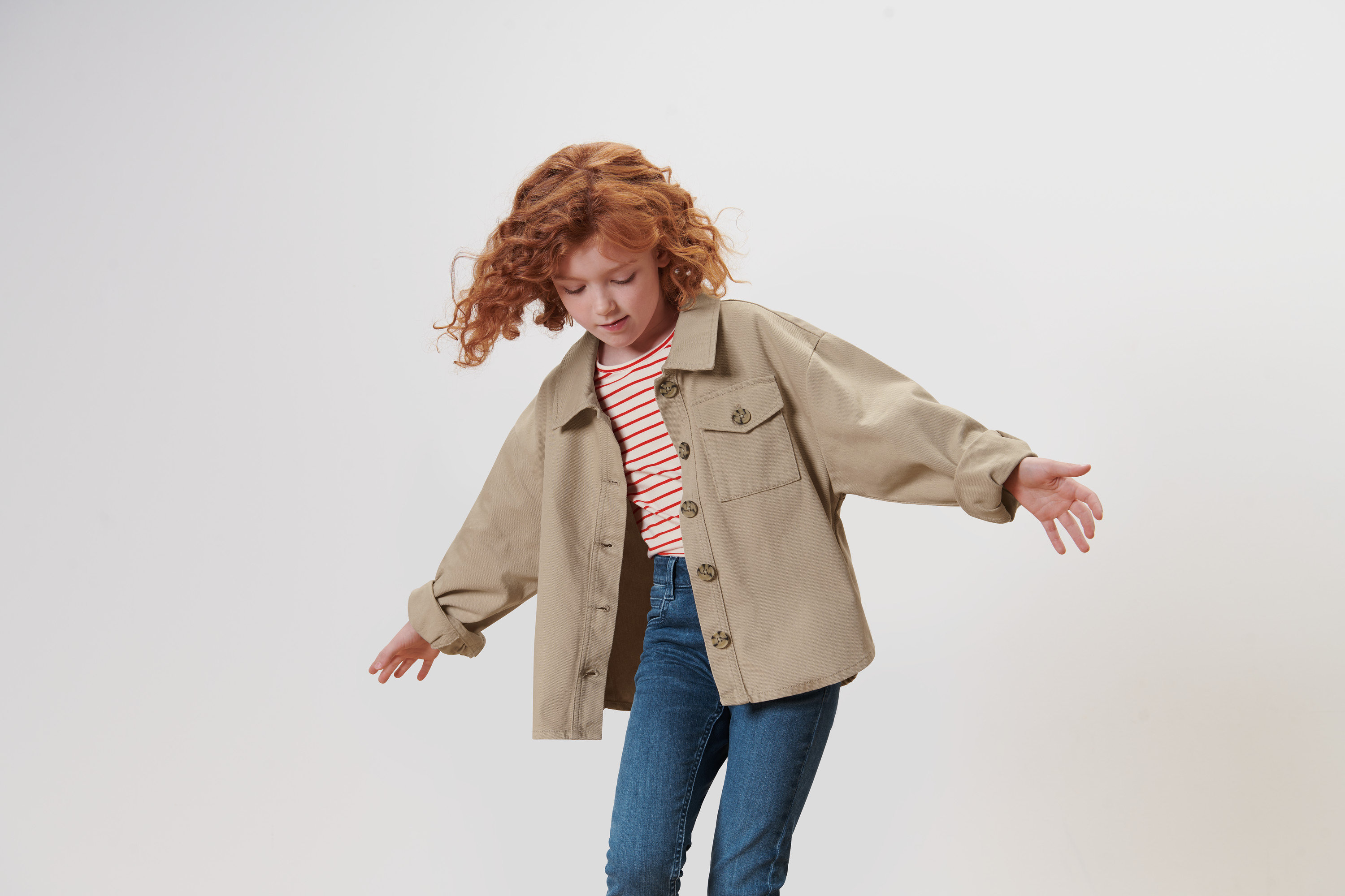 Mädchen mit lockigen roten Haaren winkt mit den Armen und trägt eine Kinder-Denimjacke.