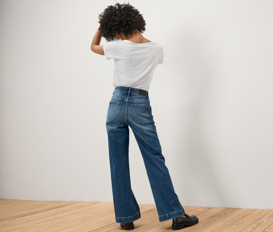 Rückansicht einer Frau mit Afro-Haar, die steht und die Hände im Haar hat. Sie trägt eine Wide Leg Jeans – Fit »Viki« und ein weißes T-Shirt.