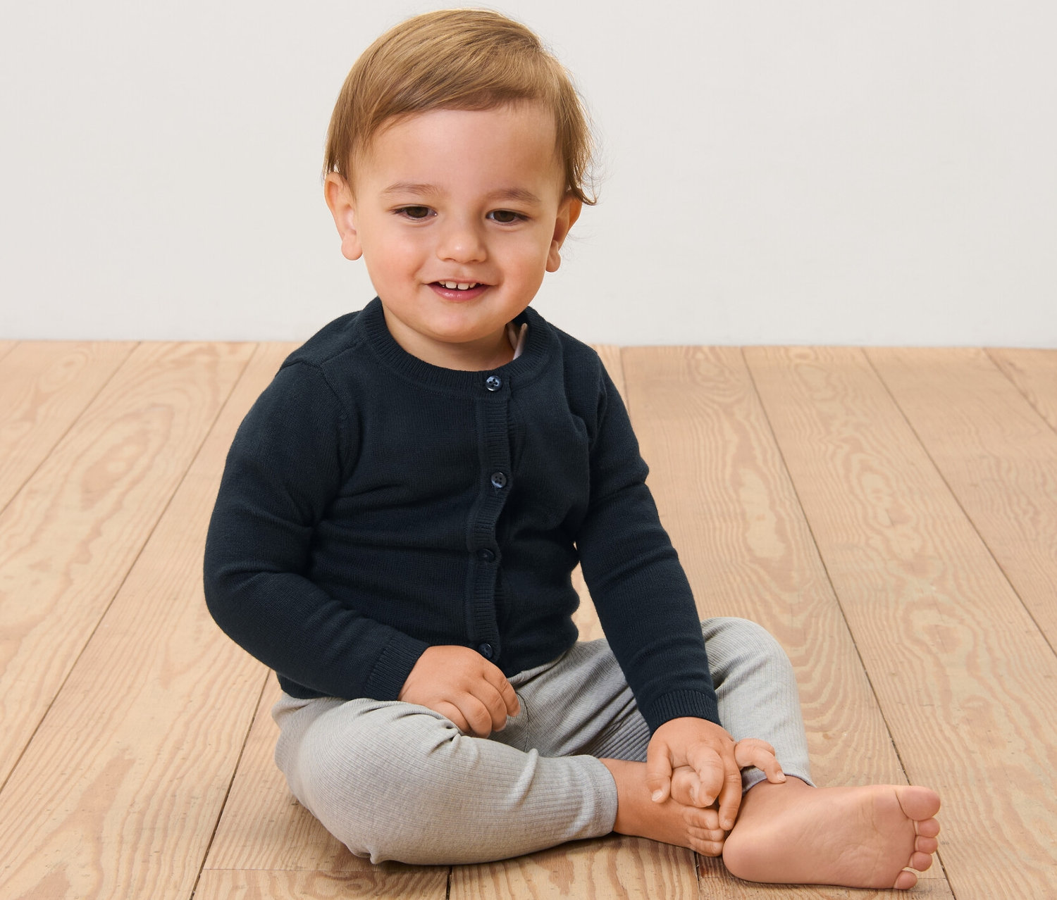 Ein Kind sitzt auf einem Holzboden und trägt eine Kinder-Strickjacke.