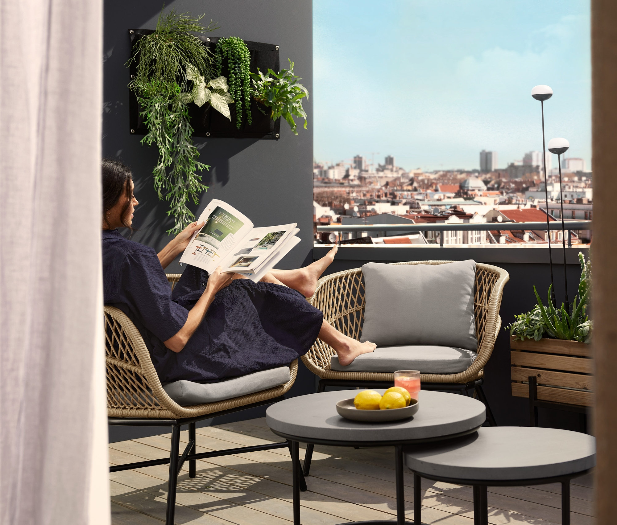 Eine Frau liest Zeitung in einem Loungesessel mit Polyrattangeflecht auf einem Balkon. In der Nähe stehen 2 Couchtische mit Beton-Tischplatte und 2 Solar-Leuchtstäbe.