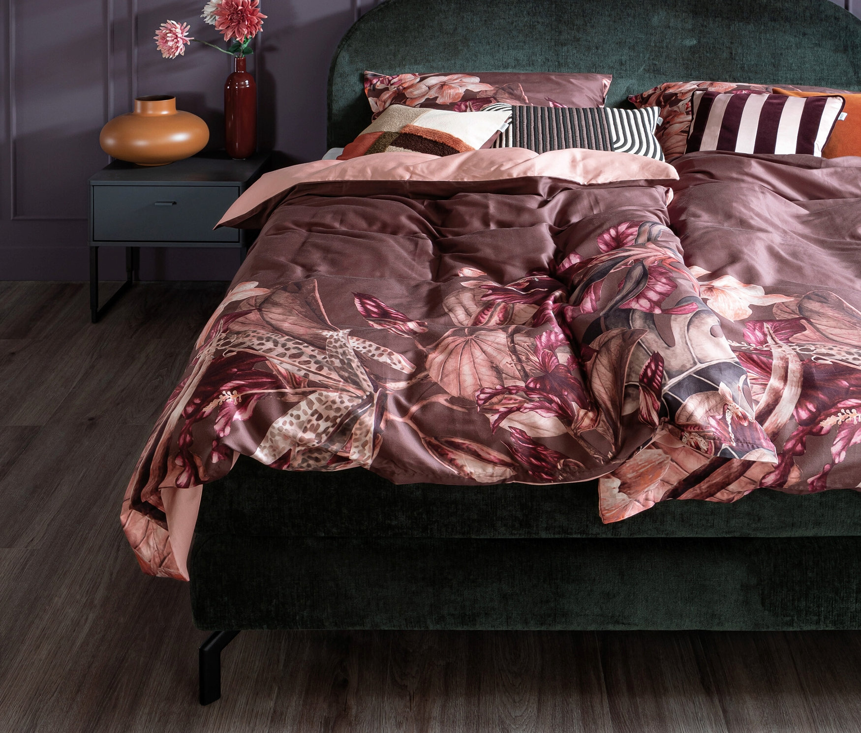 Grünes Bett mit Blumendesign-Bettwäsche und Kissen. Ein Nachttisch mit Vase und Lampe steht neben dem Bett.