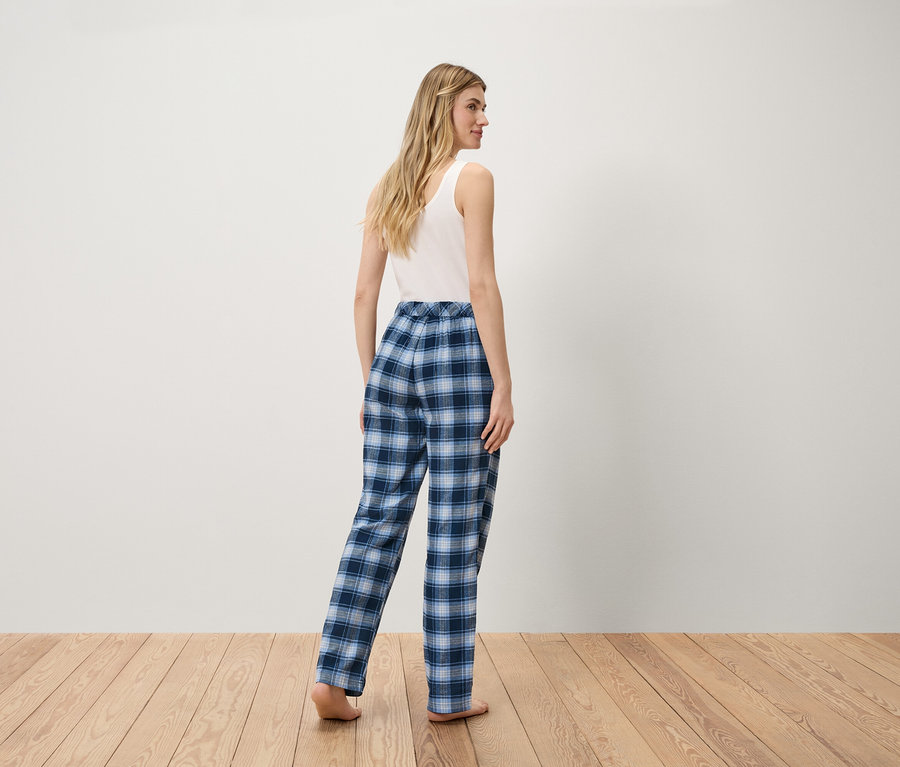 Frau in weißem Tanktop und blauer karierter Hose steht auf Holzboden.