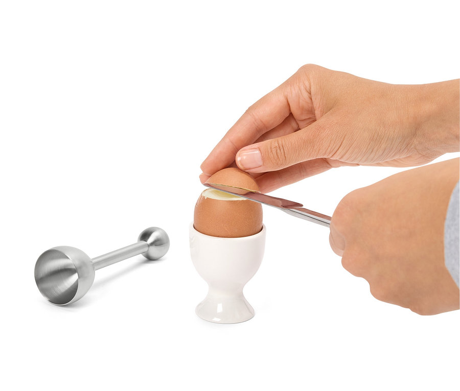 Eine Hand schneidet mit einem Messer die Spitze eines Eis in einem weißen Eierbecher ab. Links davon befindet sich ein Eierschalensollbruchstellenverursacher aus Edelstahl.