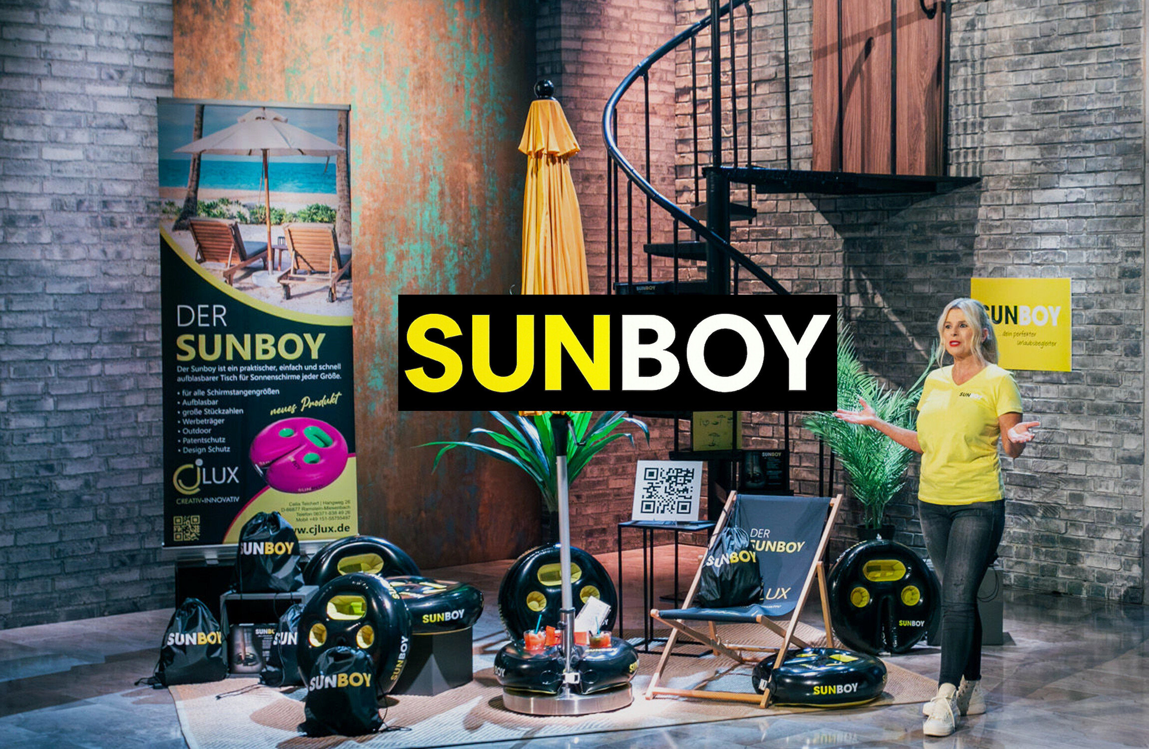 Frau präsentiert Sunboy-Produkte: aufblasbare Tische, Sonnenschirmständer, Strandstuhl und Tragetaschen vor Backsteinmauer und Wendeltreppe.