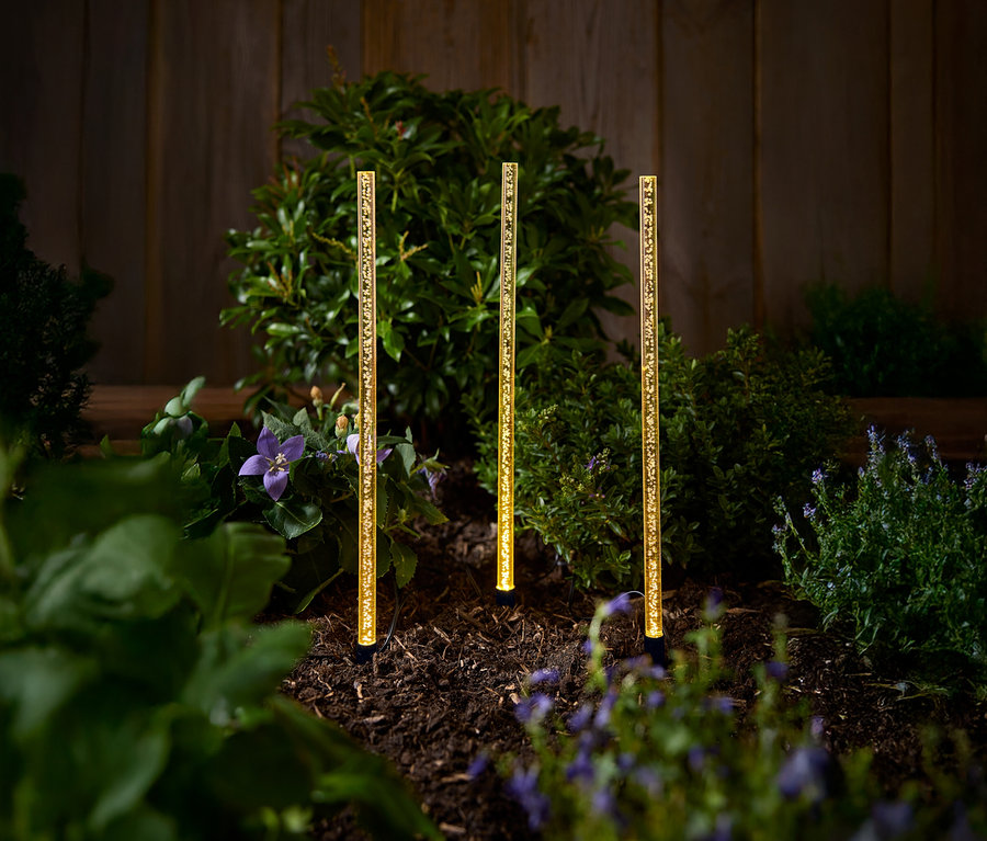 Drei goldene Solar-Gartenleuchten stehen auf einem Beet mit Büschen und Blumen.