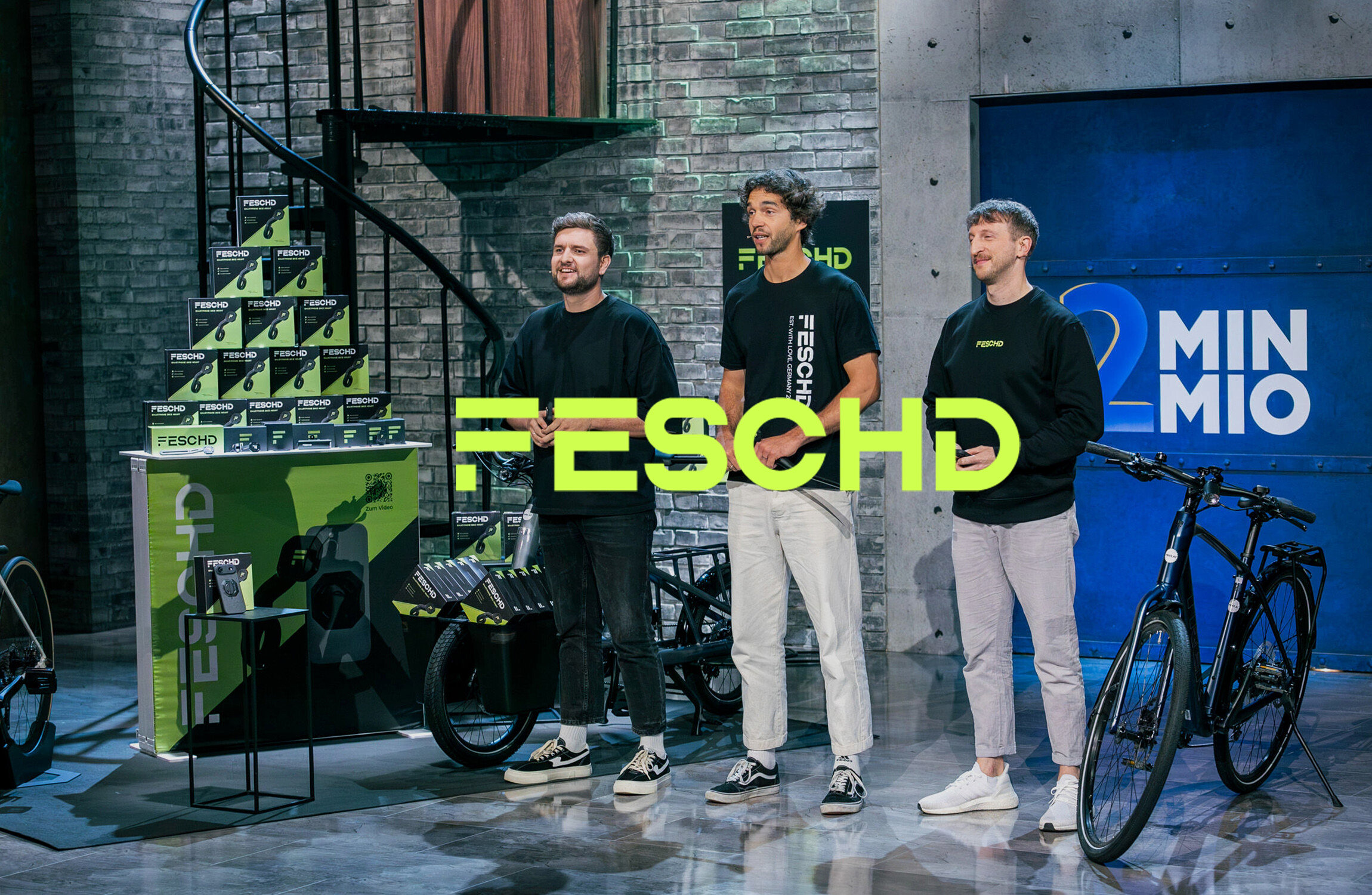 Drei Männer stehen vor einem Stand mit FESCHD-Produkten und Fahrrädern. Im Hintergrund eine Wendeltreppe und eine Wand mit der Aufschrift "2 MIN MIO".