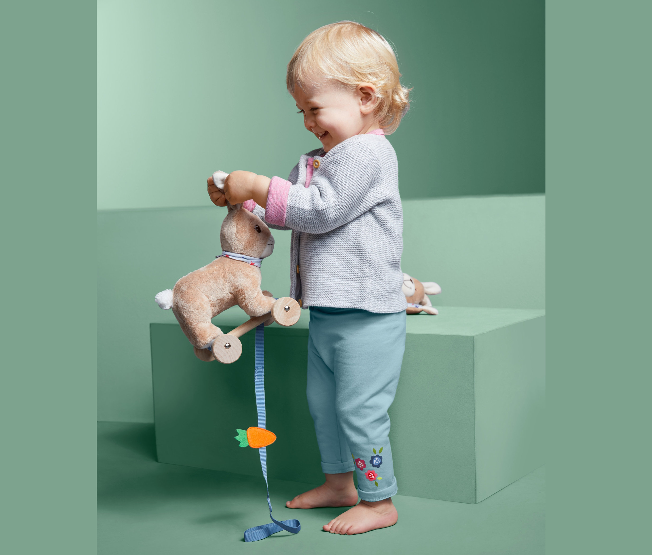 Kleinkind spielt mit Plüschhasen auf Rädern und trägt eine Sweathose.