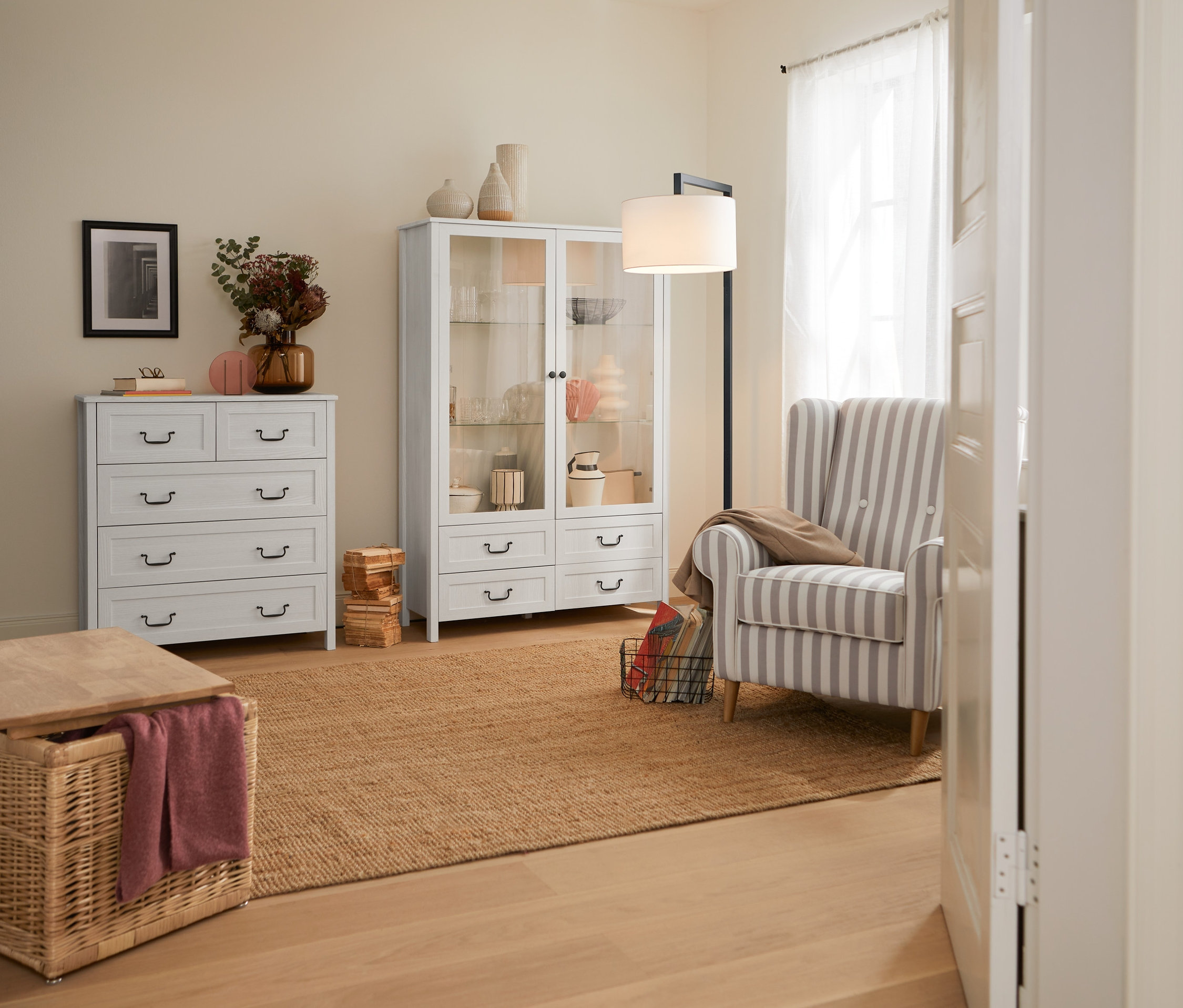 Innenaufnahme mit einem Ohrensessel »Anton« greige-weiß gestreift, einer Truhe aus Geflecht, einer Hochkommode mit 5 Schubladen, einer Vitrine, 2-türig, und einer Bogenleuchte mit Textilschirm.