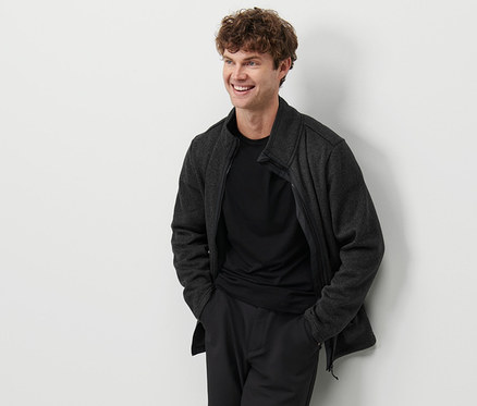 Mann an eine Wand gelehnt trägt eine anthrazitfarbene Strickfleece-Jacke.