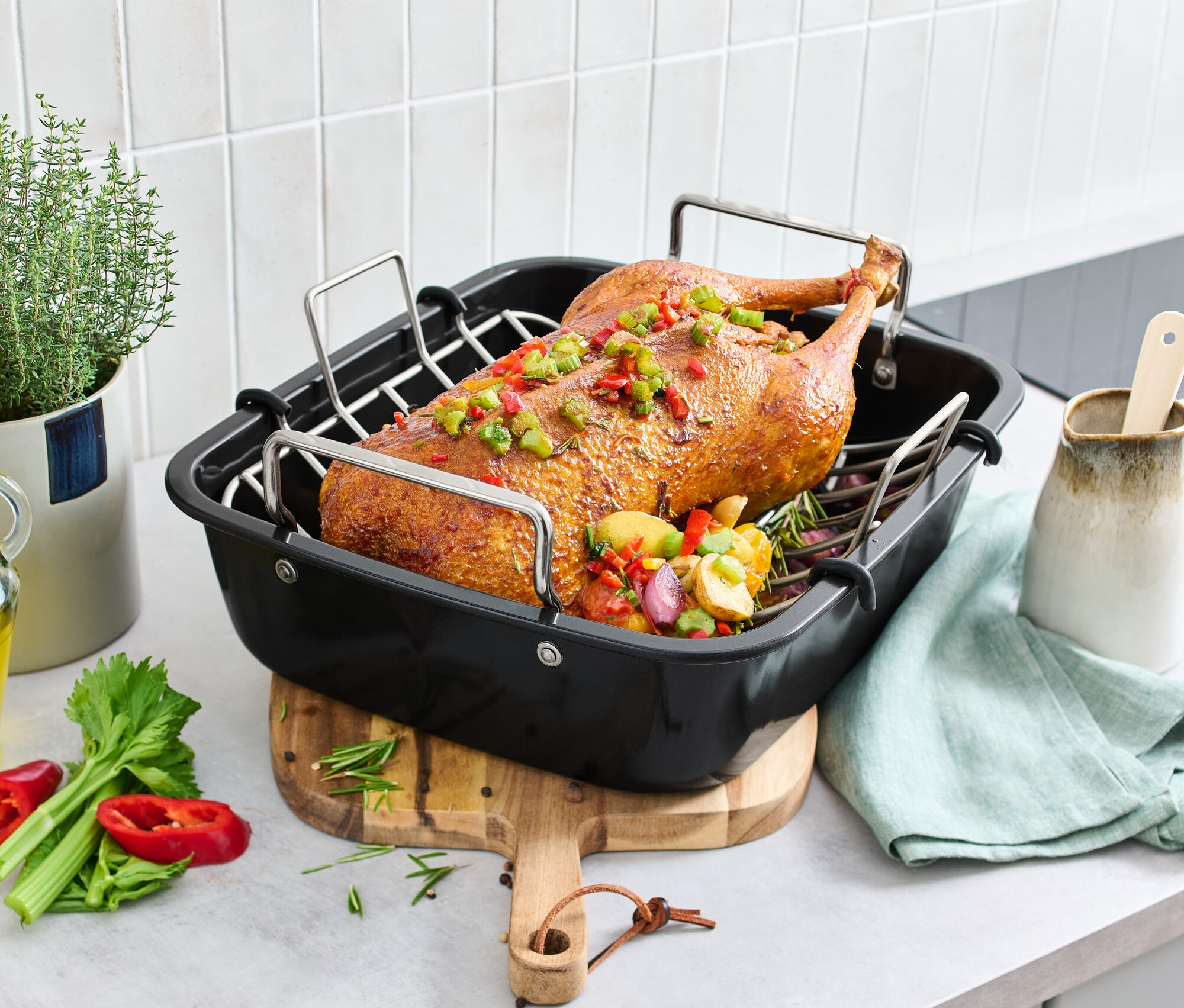 Gebratene Ente in XL-Edelstahl-Bräter mit Rost auf einem Tisch mit Gemüse und Kräutern.