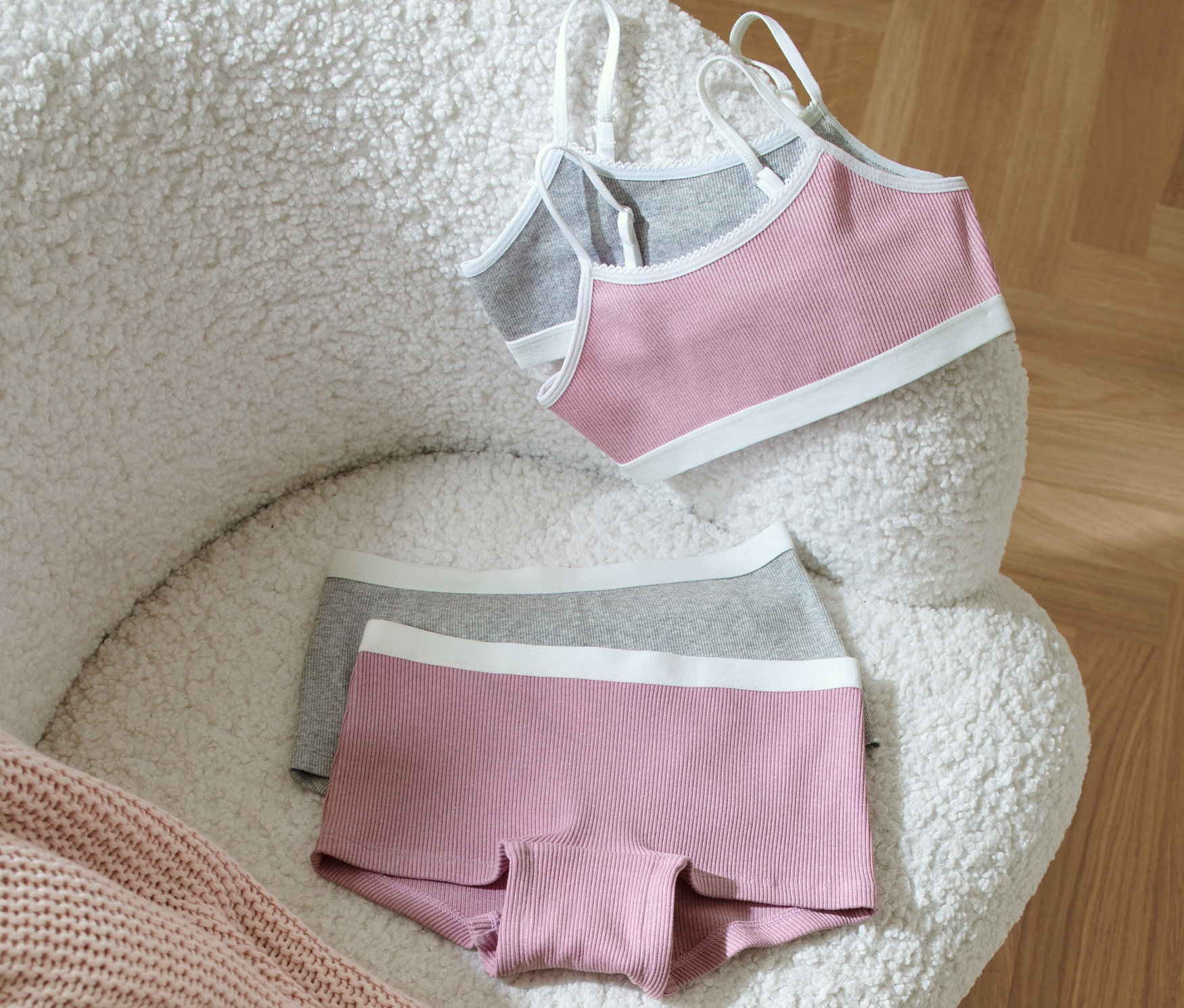 Mädchen-Wäsche-Set liegt auf einem weißen, zotteligen Sessel neben einer rosa Strickdecke.