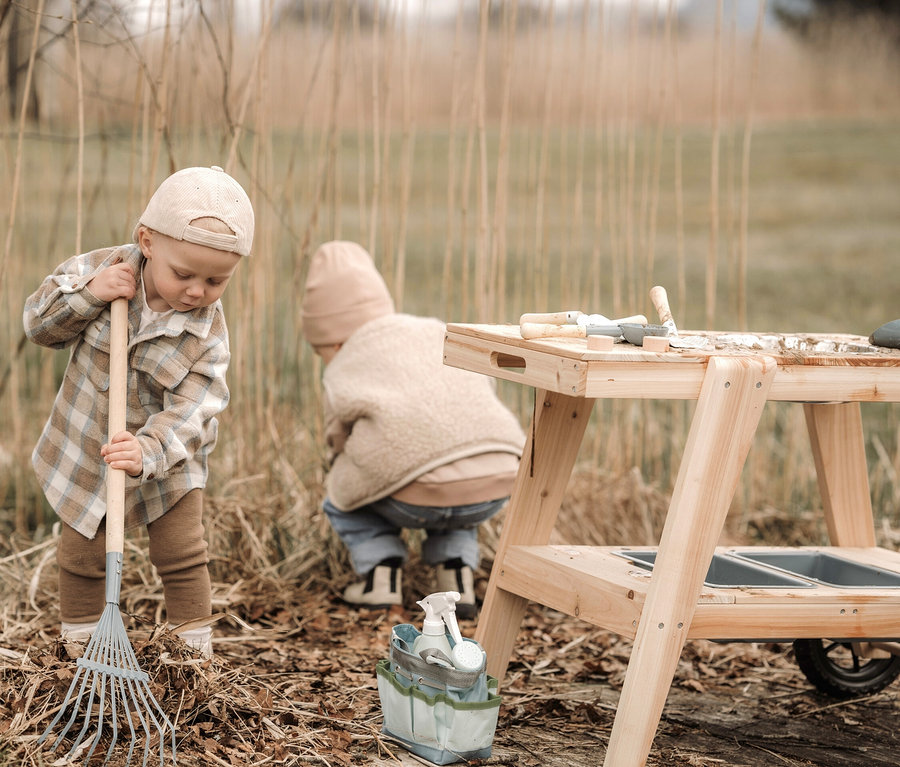 Ein Junge harkt Blätter neben einem Baum. Im Hintergrund ist ein anderes Kind an einer kompakten small foot Matschküche und einem großen small foot Gartenwerkzeug-Set.