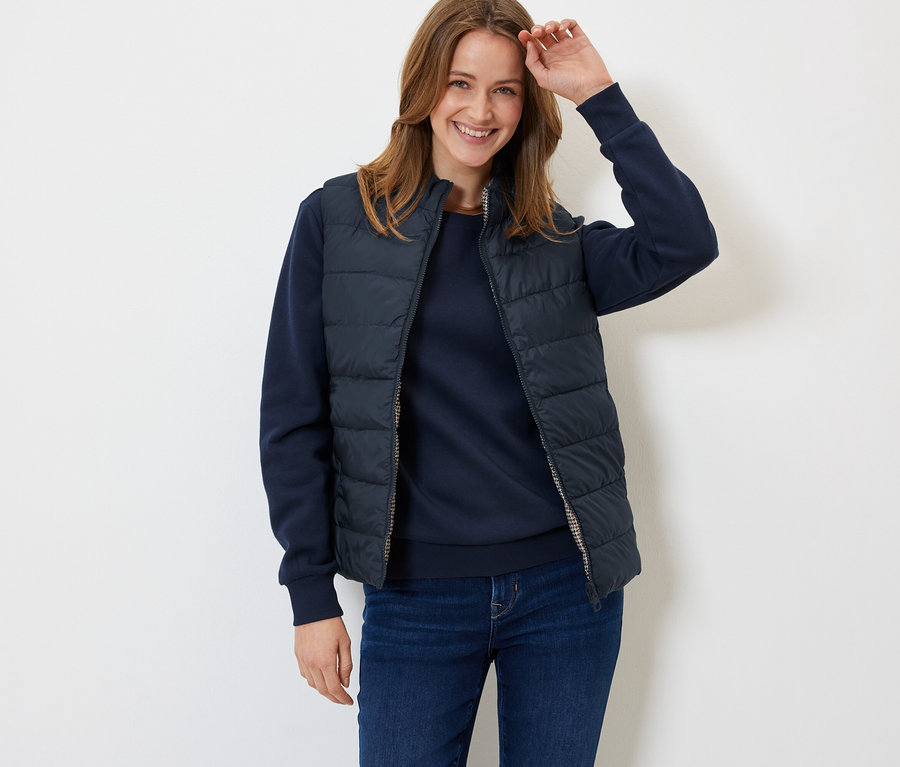 Eine lächelnde Frau posiert in einer marineblauen Steppweste, einem Pullover und Jeans.