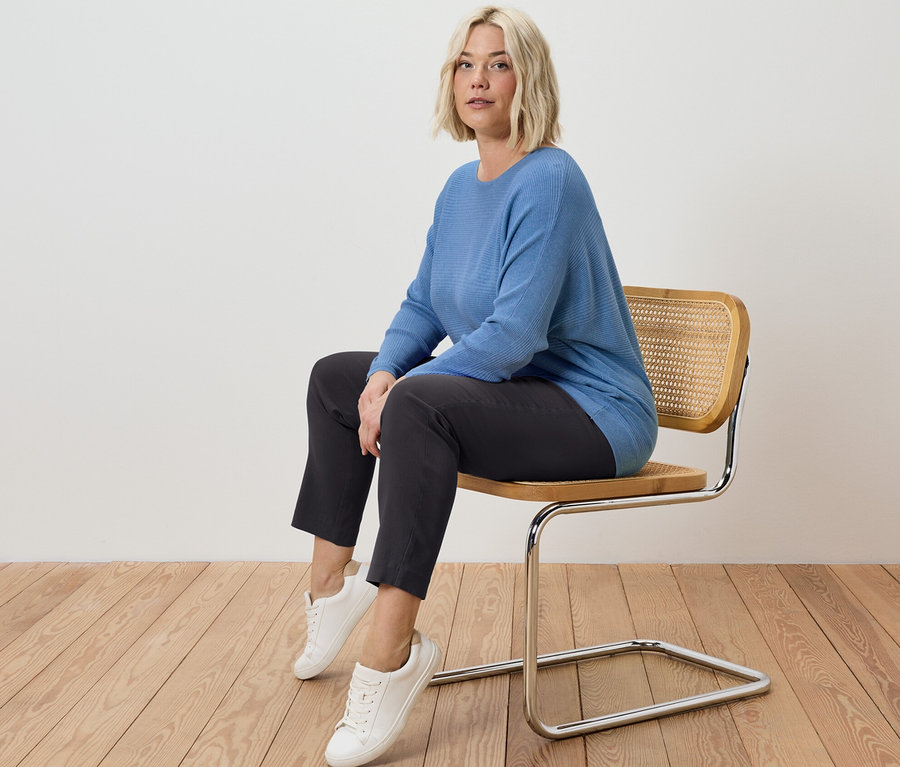 Blonde Frau sitzt auf einem Stuhl und trägt eine Stretchhose und einen blauen Feinstrickpullover.