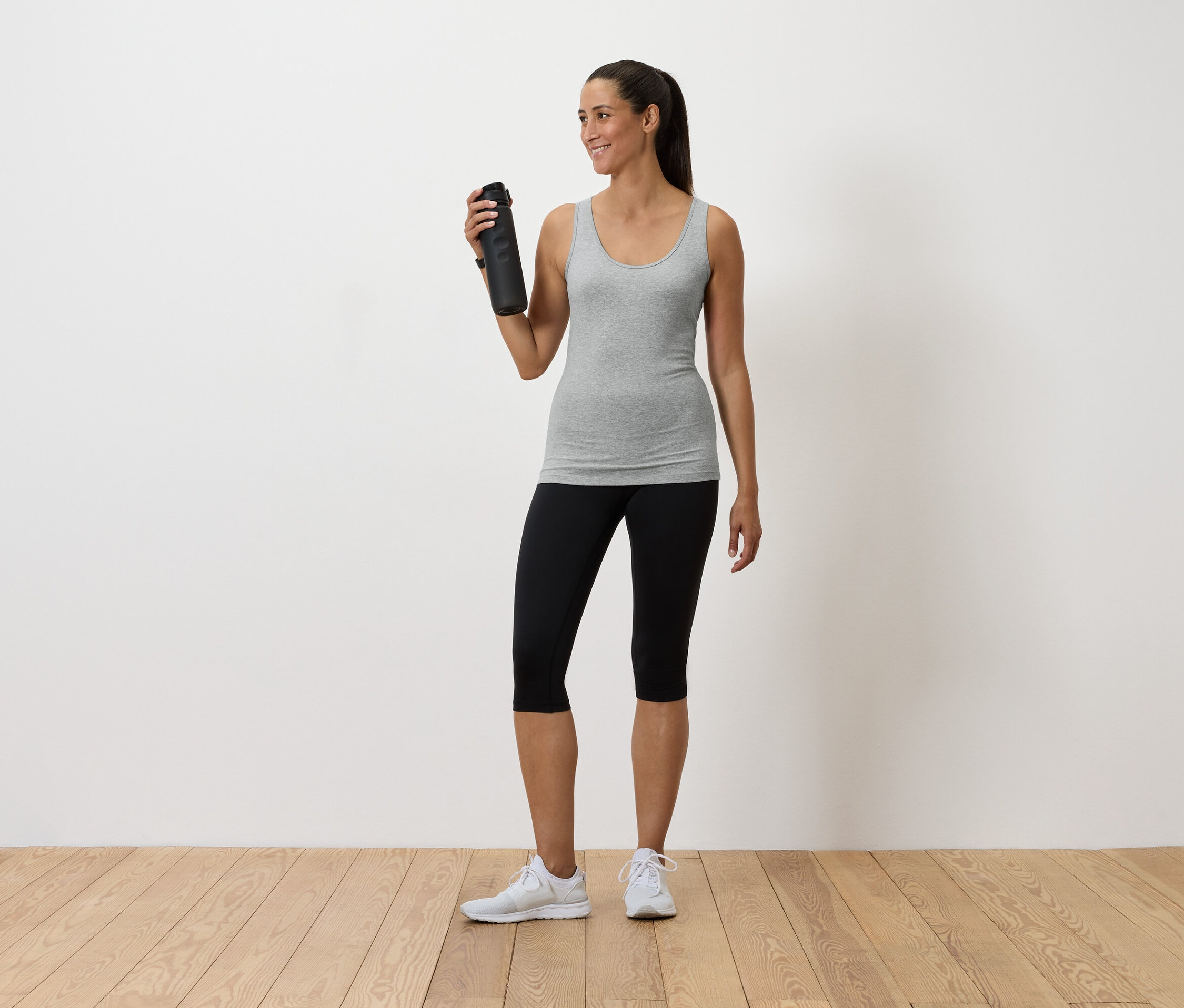 Frau hält eine Wasserflasche, trägt ein graues Tanktop, schwarze Leggings und weiße Turnschuhe.