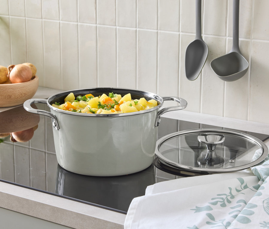 Auf einem Kochfeld steht ein Topf mit Deckel und Gemüse. Im Hintergrund befinden sich eine Schüssel mit Kartoffeln und aufgehängte Schöpfkellen.