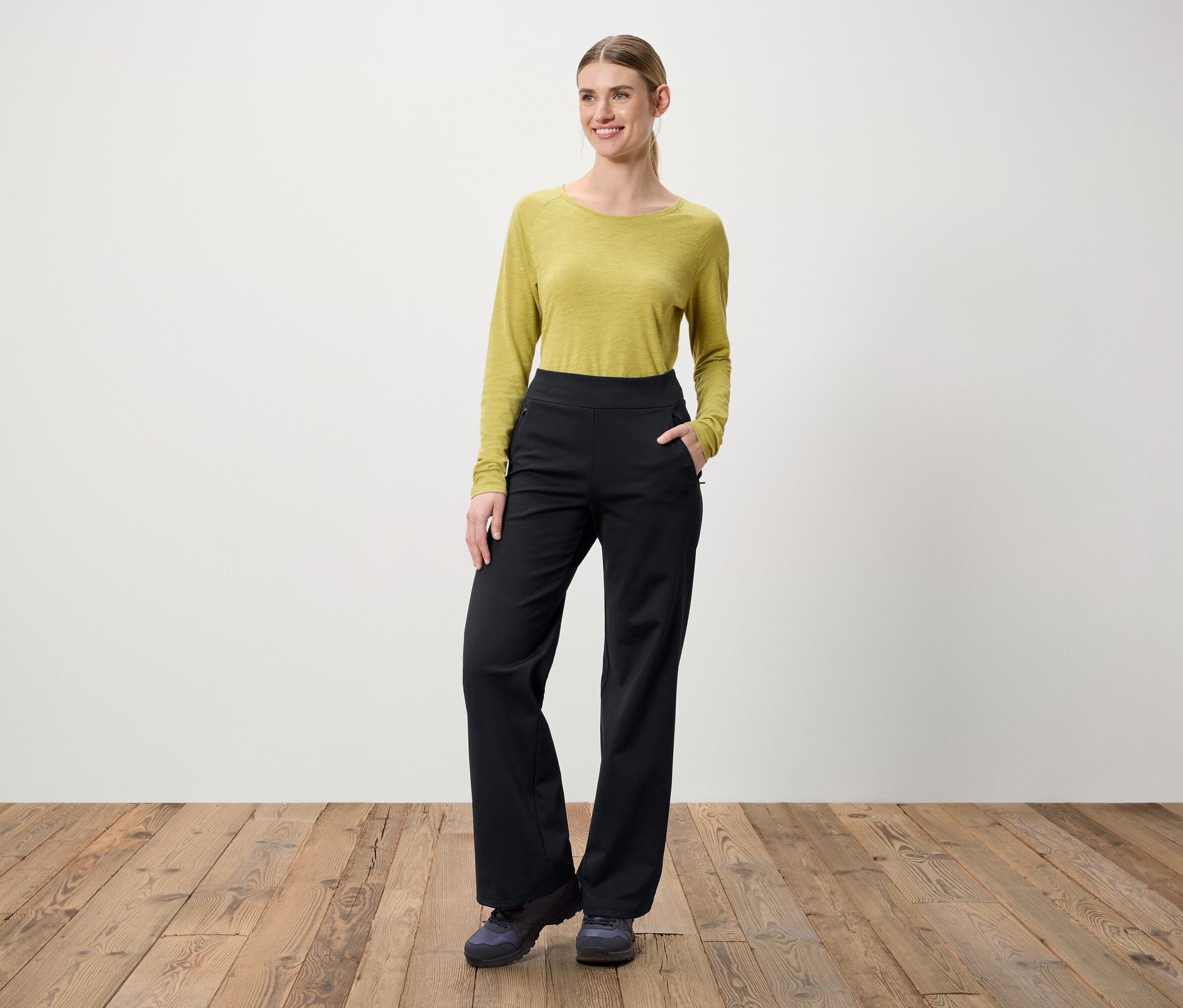 Eine Frau steht auf einem Holzboden. Sie trägt eine grüne Bluse mit langen Ärmeln und eine schwarze Hose.