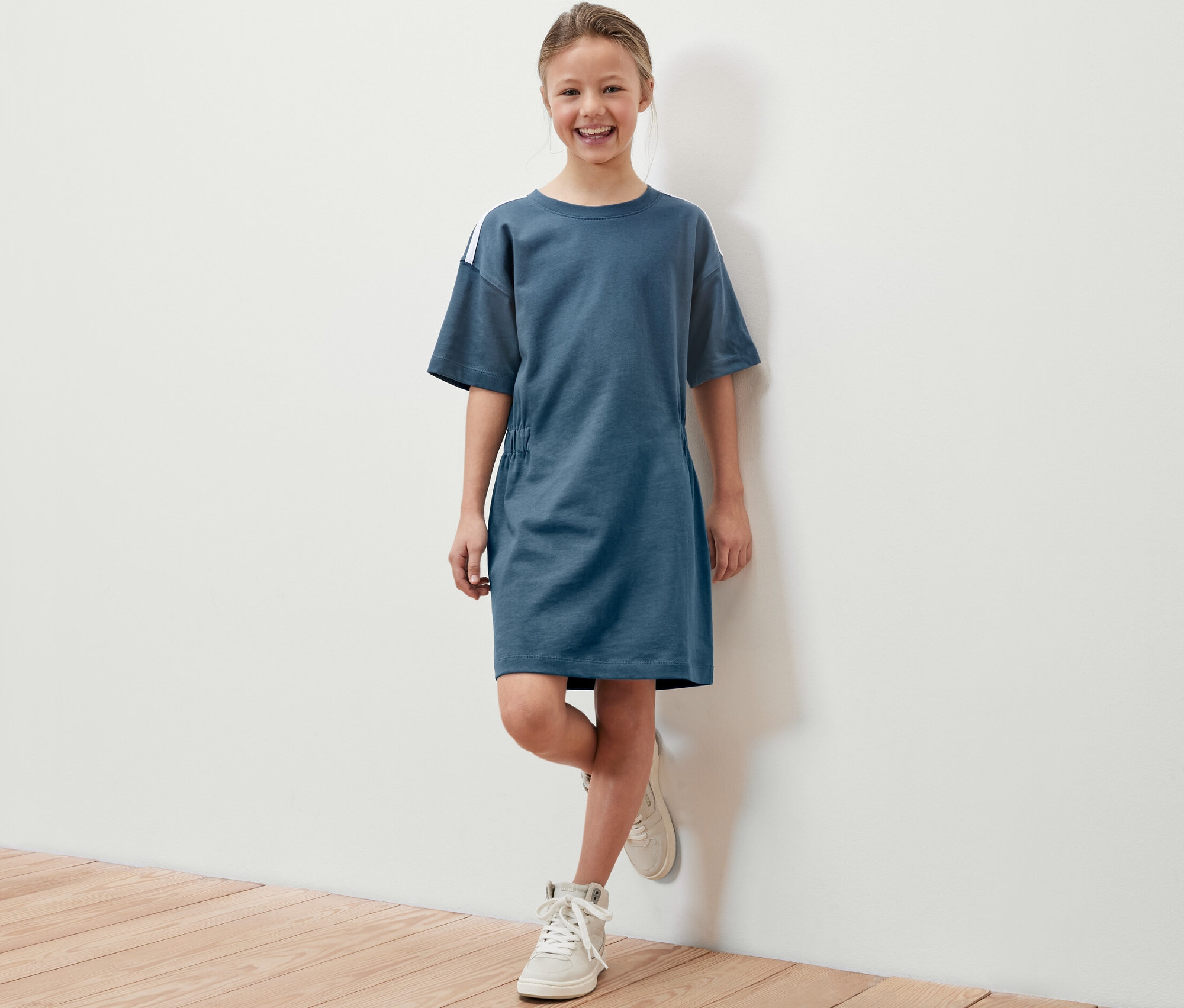 Lächelndes Mädchen posiert in blauem Kinder-Heavy-Jersey-Kleid mit weißen Schuhen.