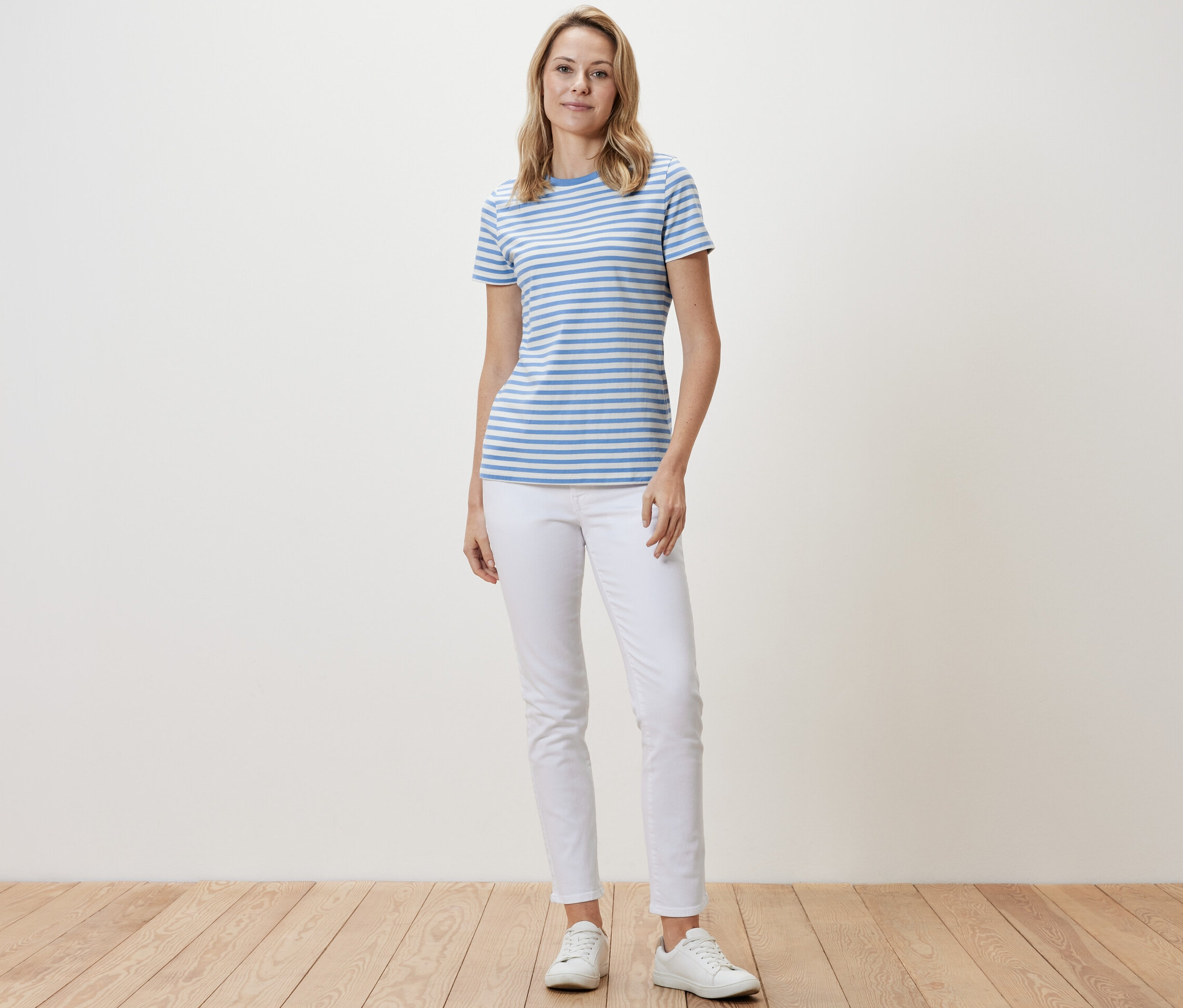 Eine Frau steht und trägt ein Basic T-Shirt, hellblau-weiß gestreift, weiße Hosen und weiße Turnschuhe.