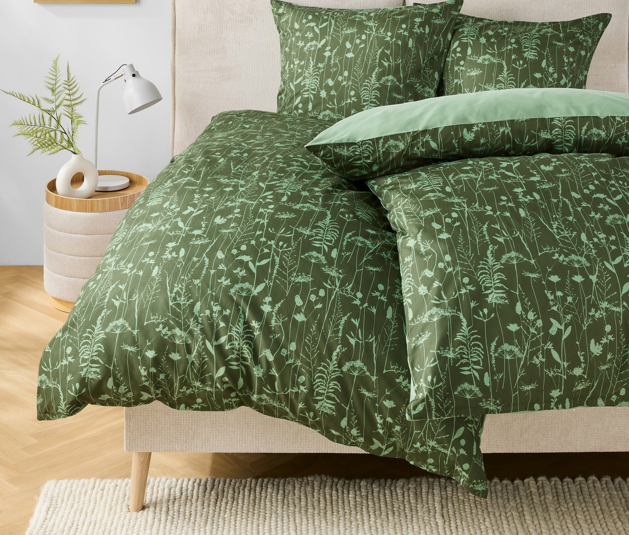 Grüne Bettwäsche mit Blumenmuster auf einem Bett. Auf dem Nachttisch stehen eine Lampe und eine Vase mit einer Pflanze.