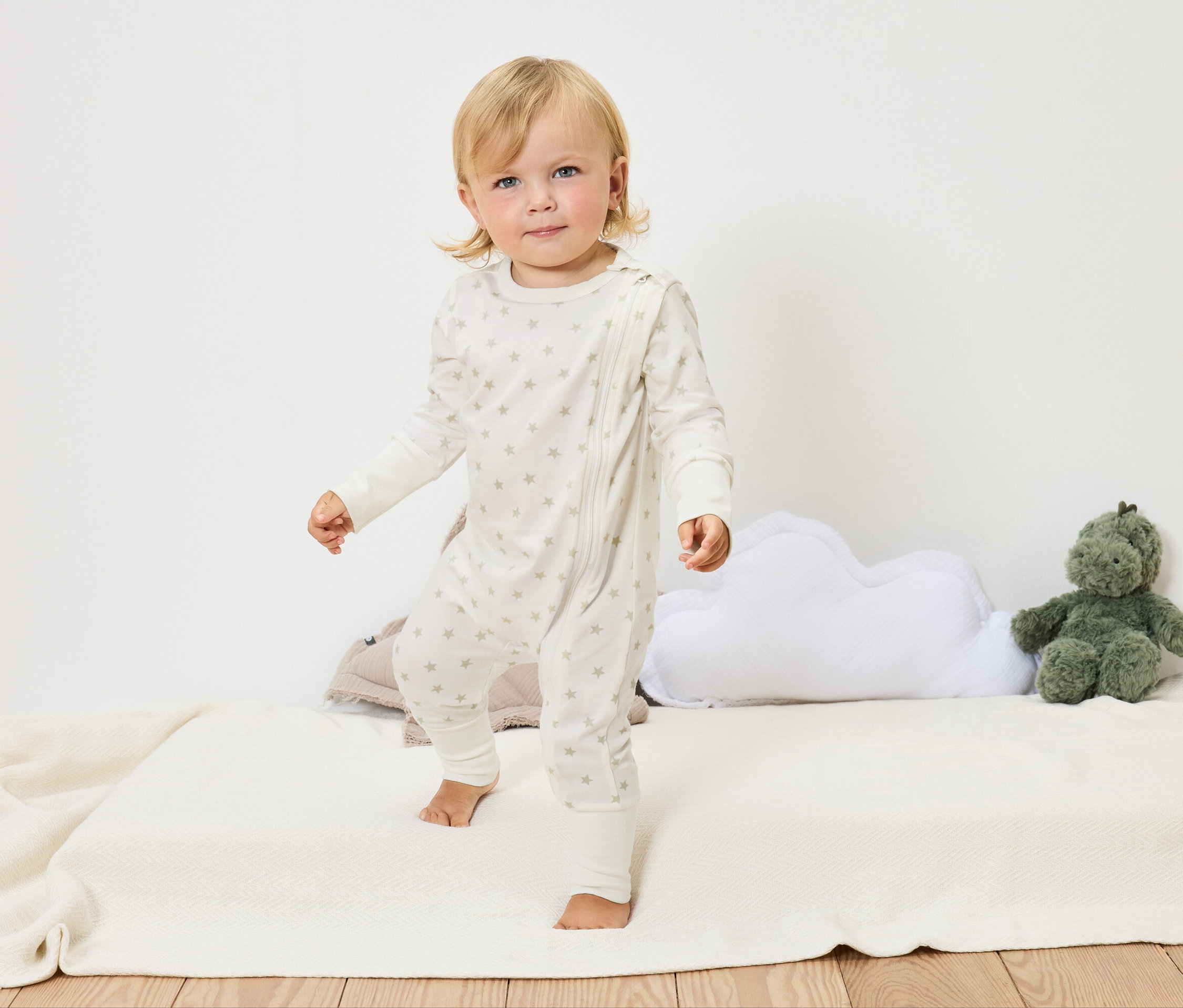 Ein blondes Kleinkind in einem weißen Sternenoverall steht barfuß auf einer weißen Decke neben einem Wolkenkissen und einem grünen Stofftier.