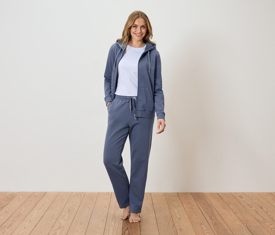 Frau steht barfuß auf Holzboden und trägt einen blauen Trainingsanzug mit Kapuzenjacke über einem weißen T-Shirt.