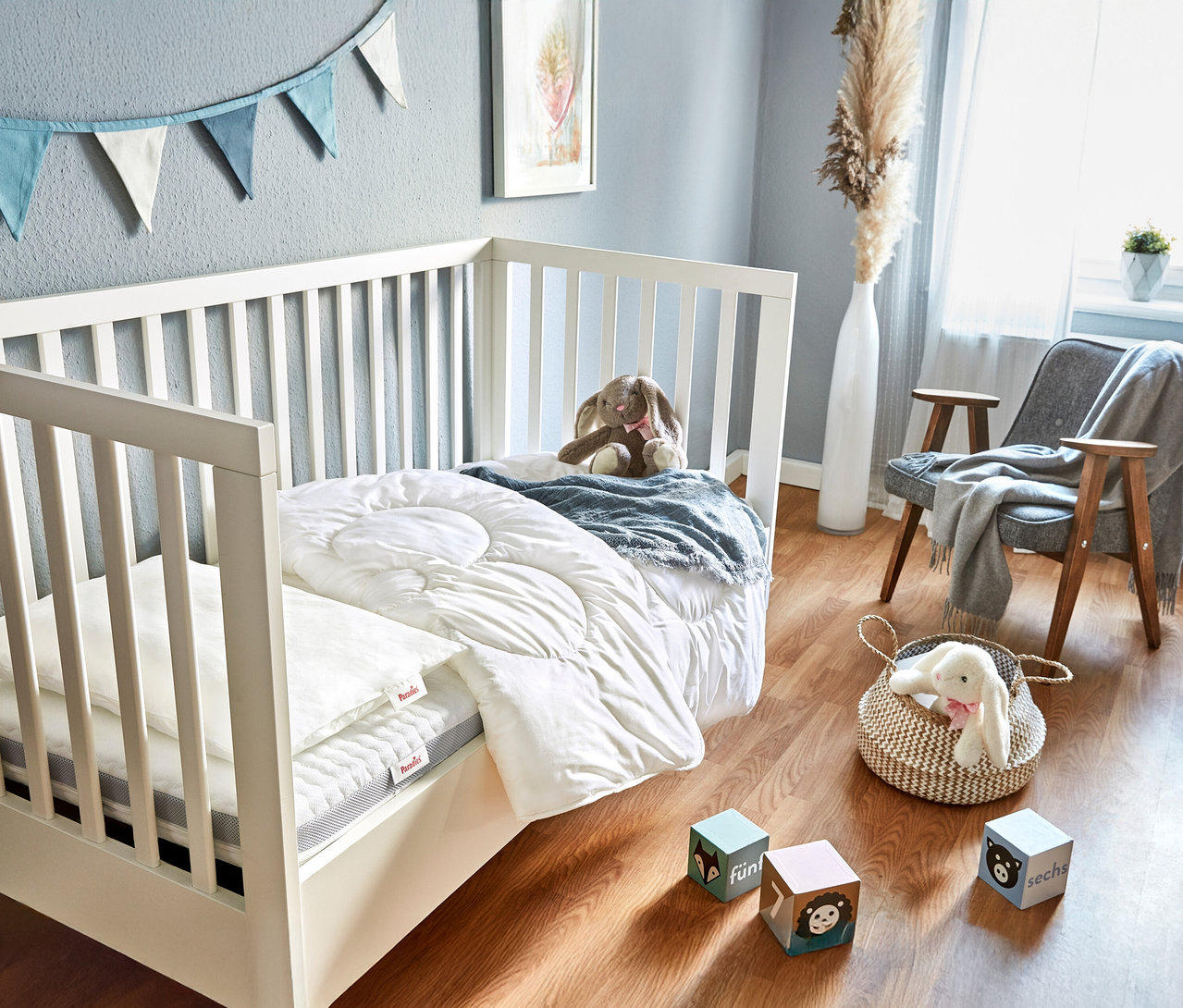 Weißes Kinderbett mit Paradies Baby- und Kindermatratze »Traumwolke«, Stofftier und blau-weißer Wimpelkette an grauer Wand.