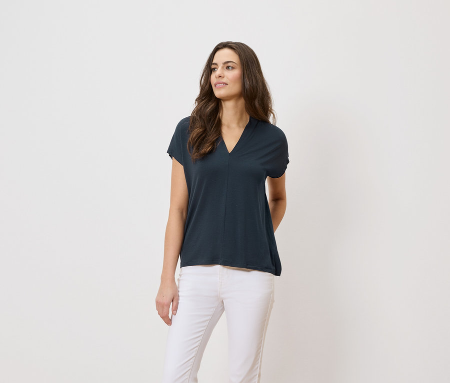 Frau trägt ein dunkelblaues T-Shirt und weiße Hosen vor einem weißen Hintergrund.