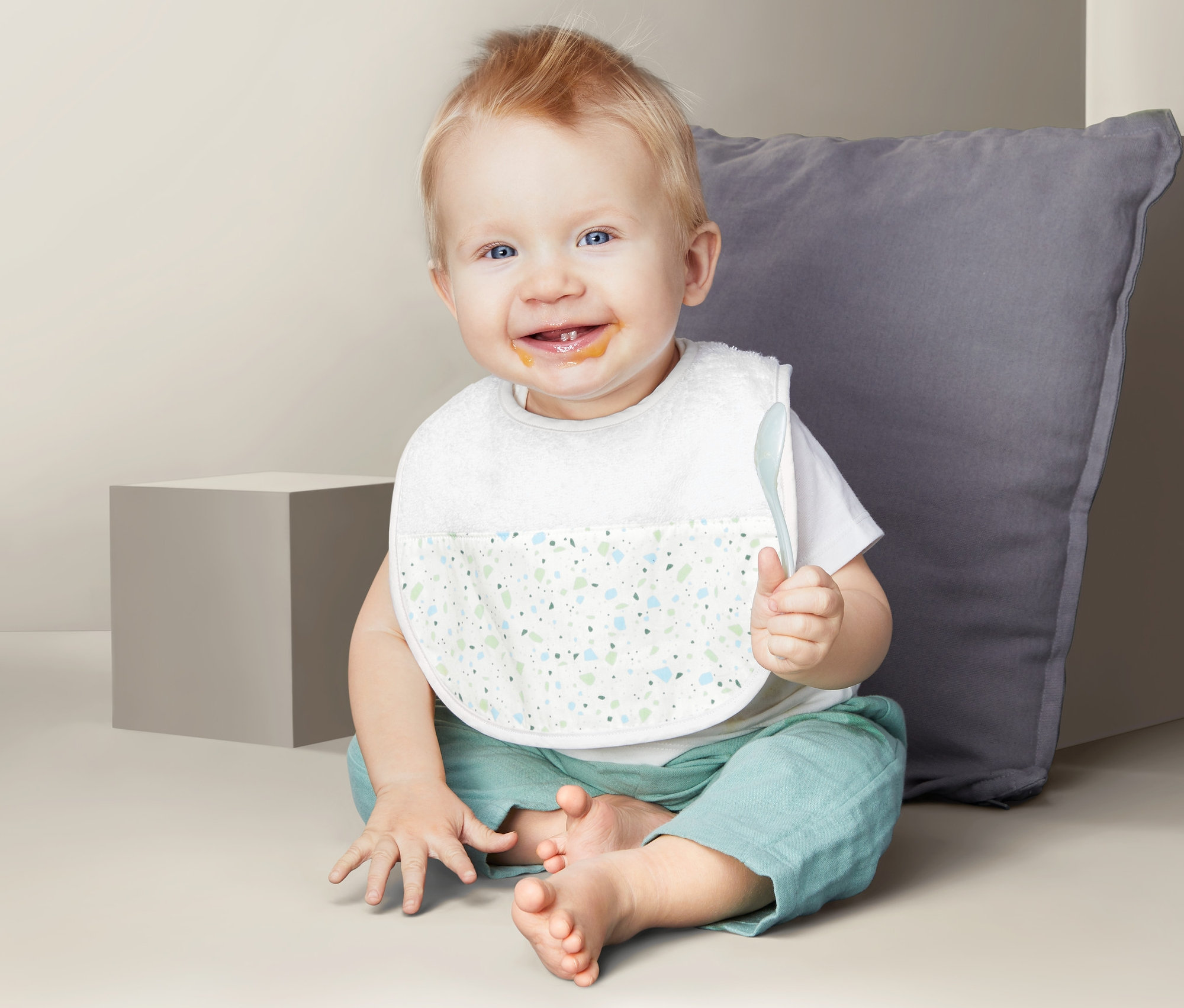 Lächelnder Säugling sitzt mit 2 Baby-Lätzchen und einem Löffel in der Hand auf dem Boden.