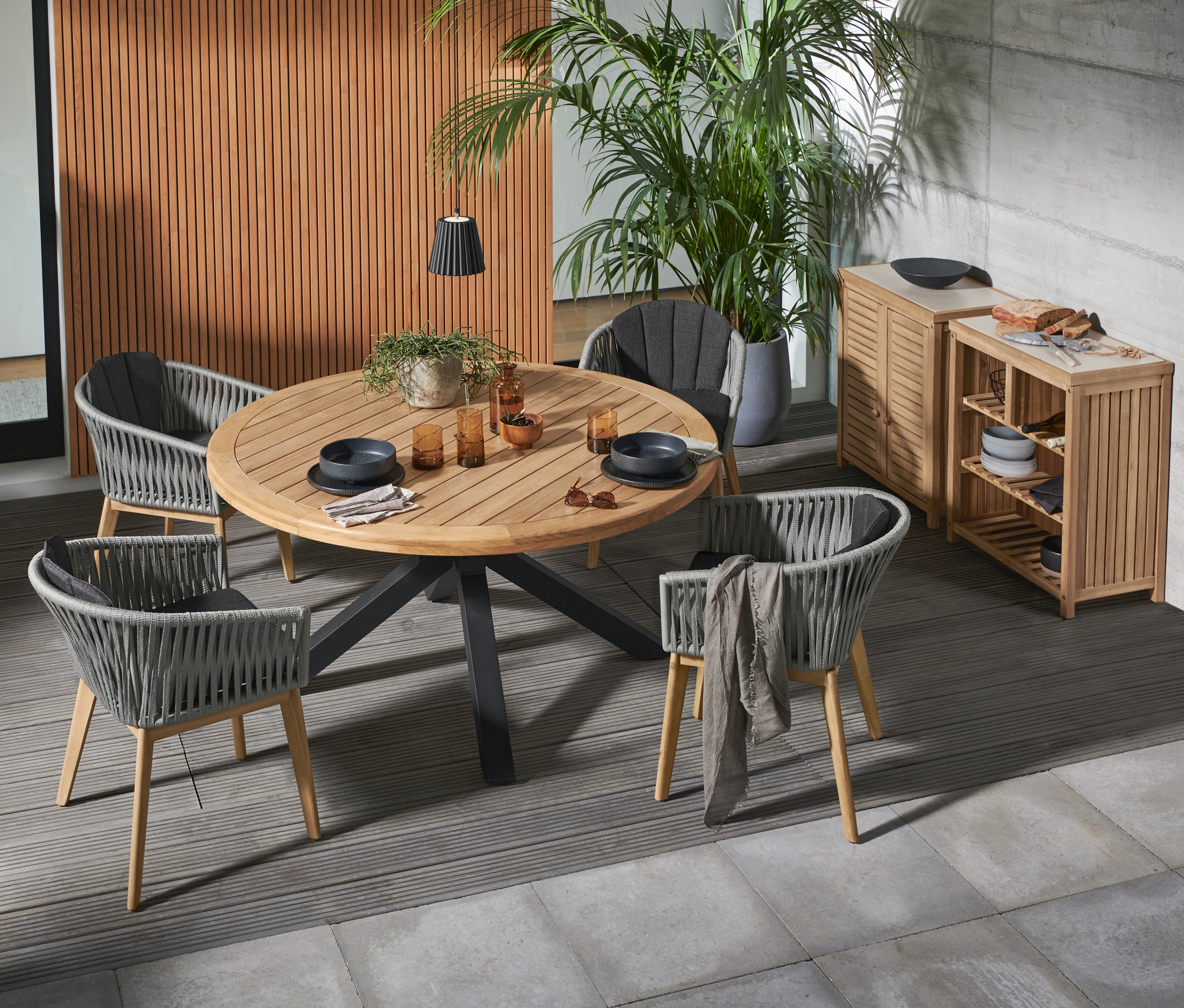 Vier Dining-Sessel mit Textilgeflecht umgeben einen Premium-Gartentisch aus Teakholz. Im Hintergrund befinden sich ein Outdoor-Schrank »Frieso« mit Beton-Arbeitsfläche und ein offener Outdoor-Schrank »Frieso« mit Beton-Arbeitsfläche.