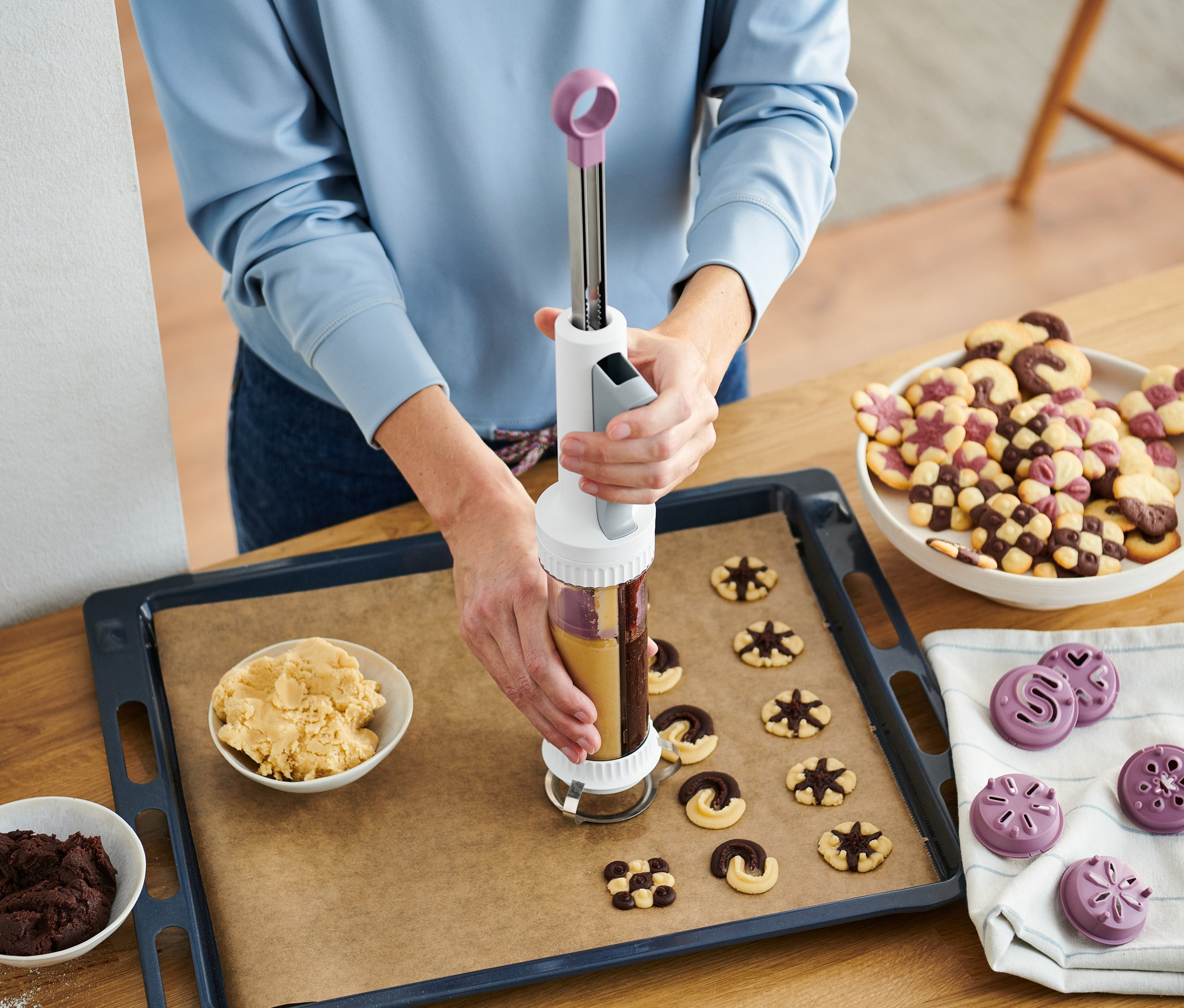 Frau verwendet Kekspresse, um Teig auf ein Backblech zu drücken. Schüsseln mit Teig und fertige Kekse auf dem Tisch.