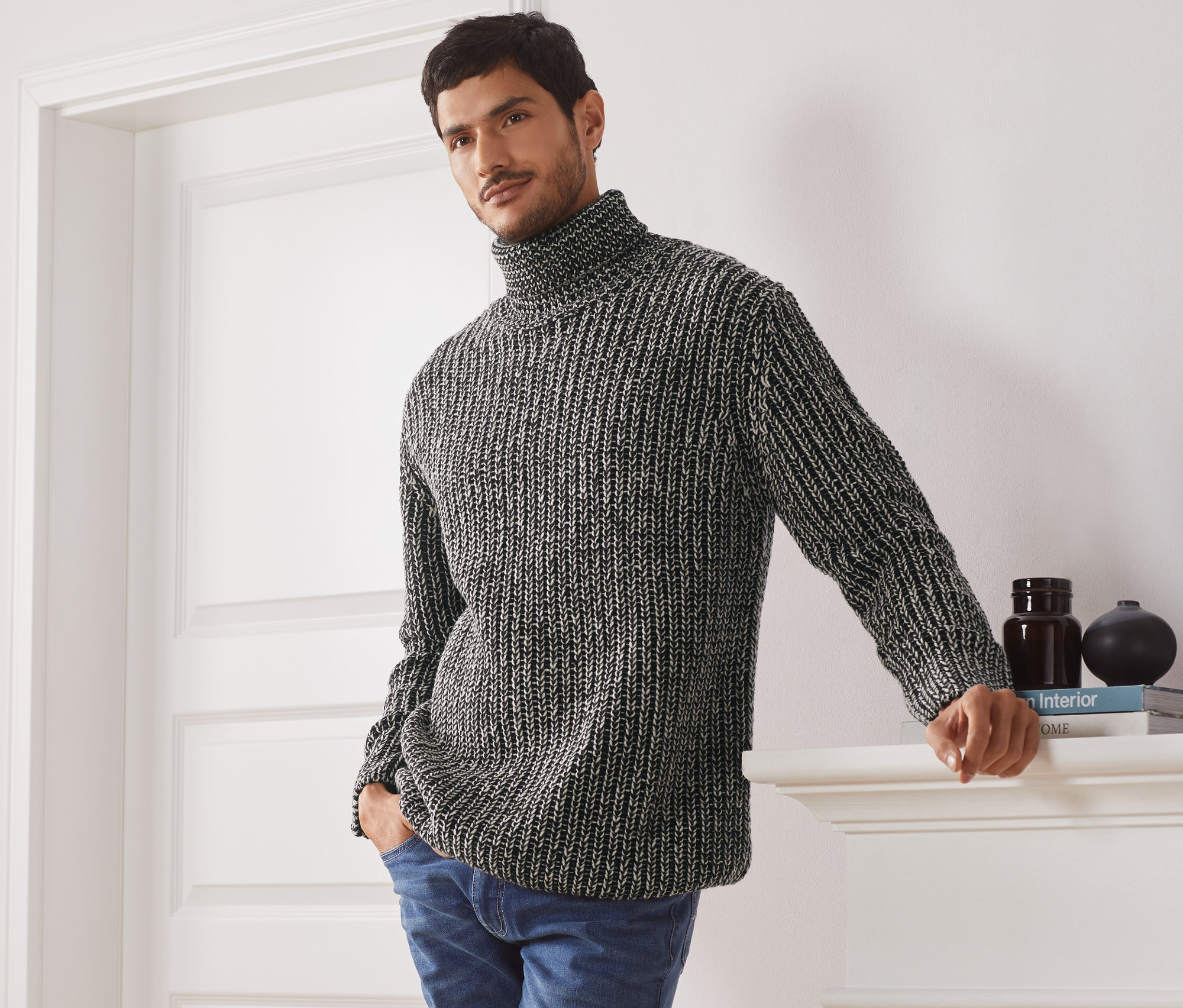 Mann posiert in Grobstrickpullover mit Rollkragen und Jeans.